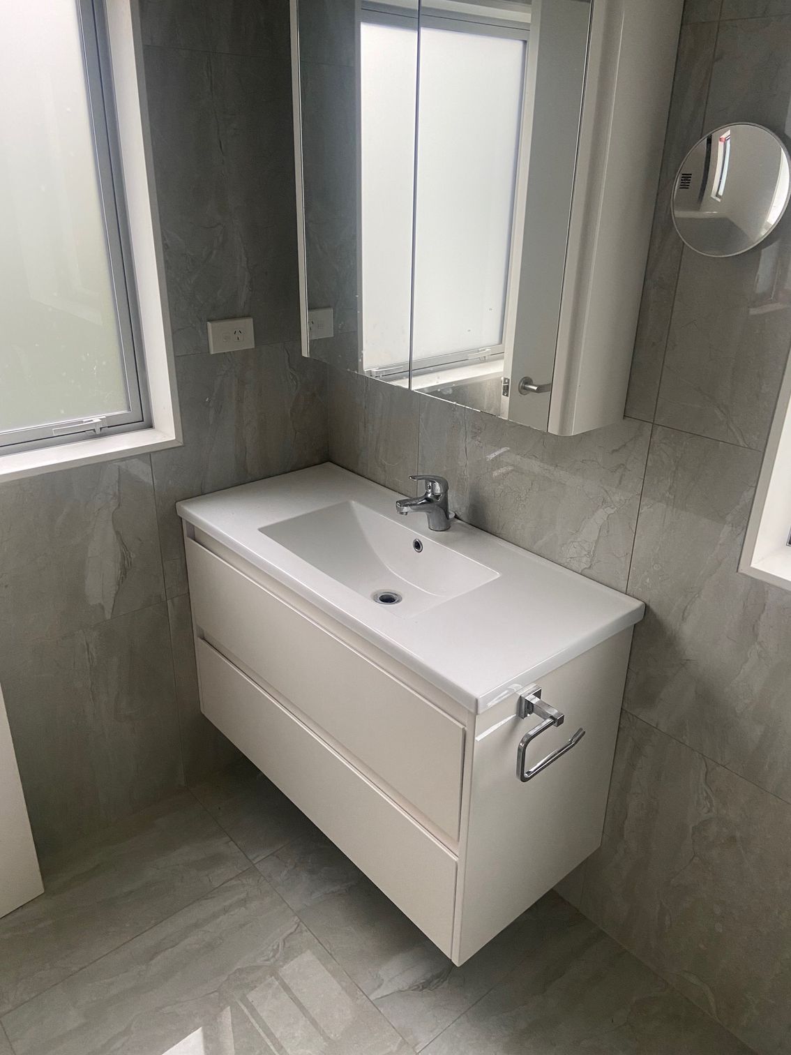 Modern white bathroom vanity with sink, mirror, and chrome fixtures, set against gray tiled walls.
