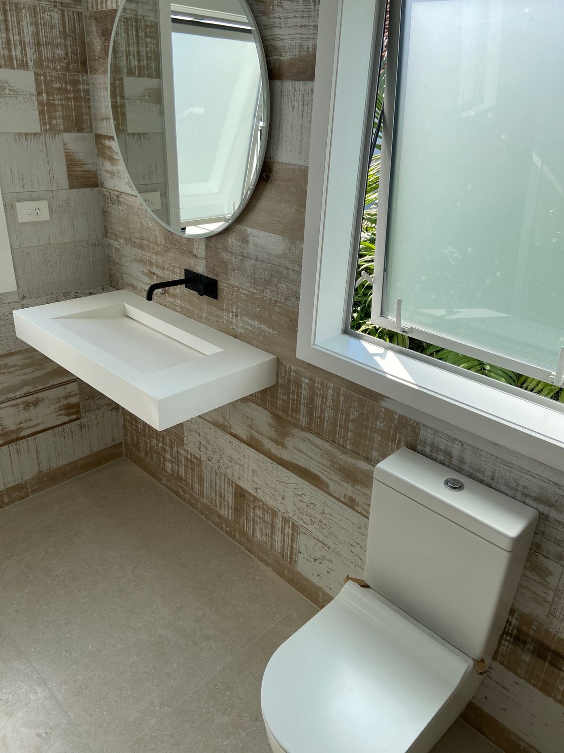 Bathroom with a white floating sink, oval mirror, and toilet, next to a window.