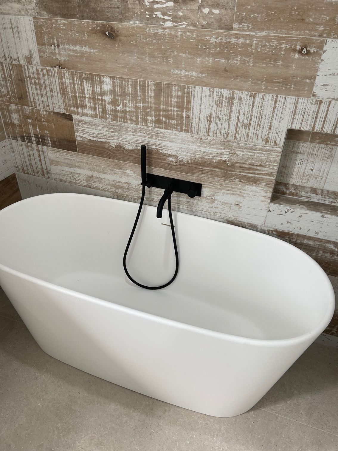 A white oval bathtub with black faucet and shower head against a wood-look tiled wall.