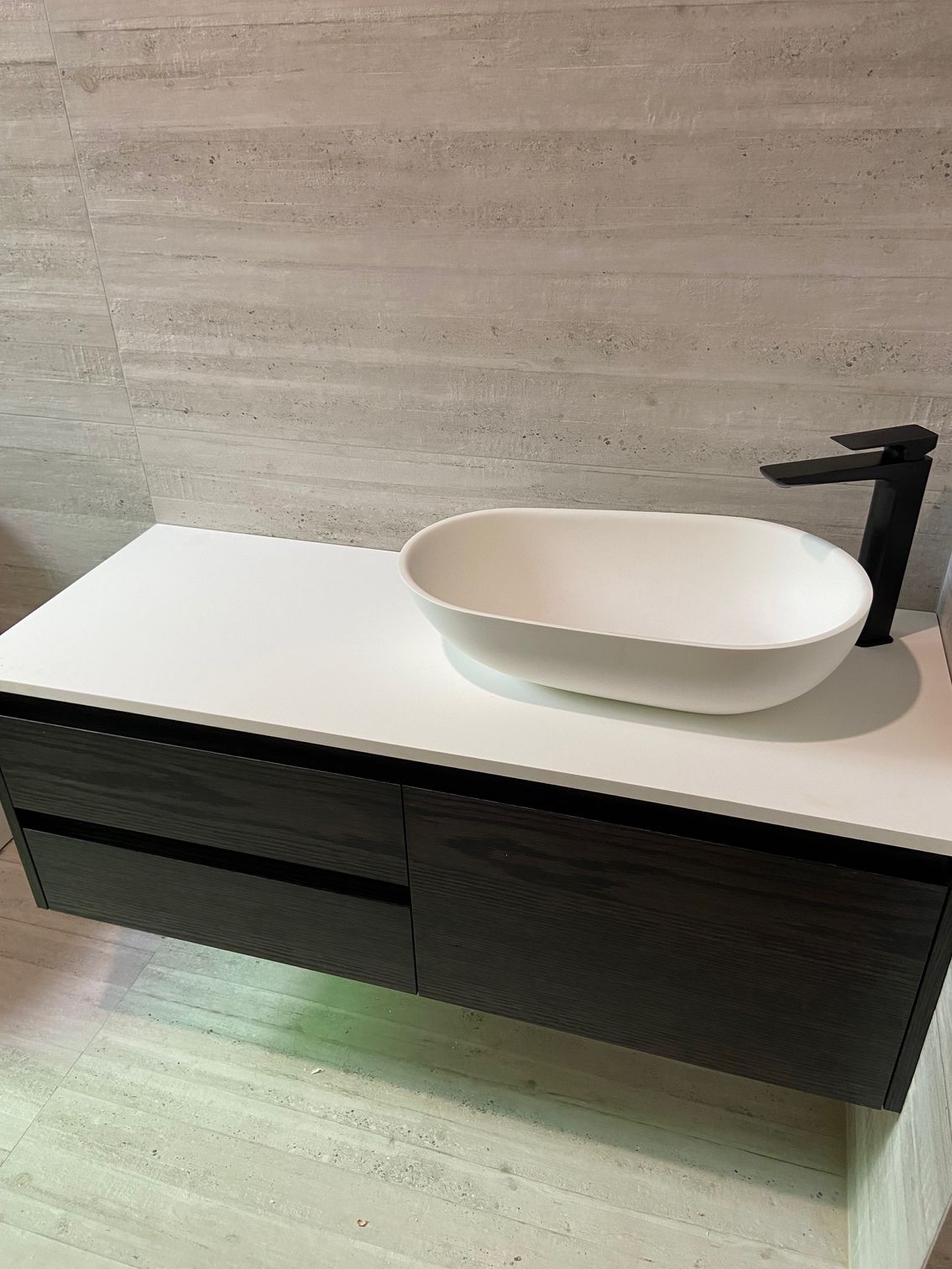 Dark vanity with white countertop, oval sink, and black faucet against a textured wall.