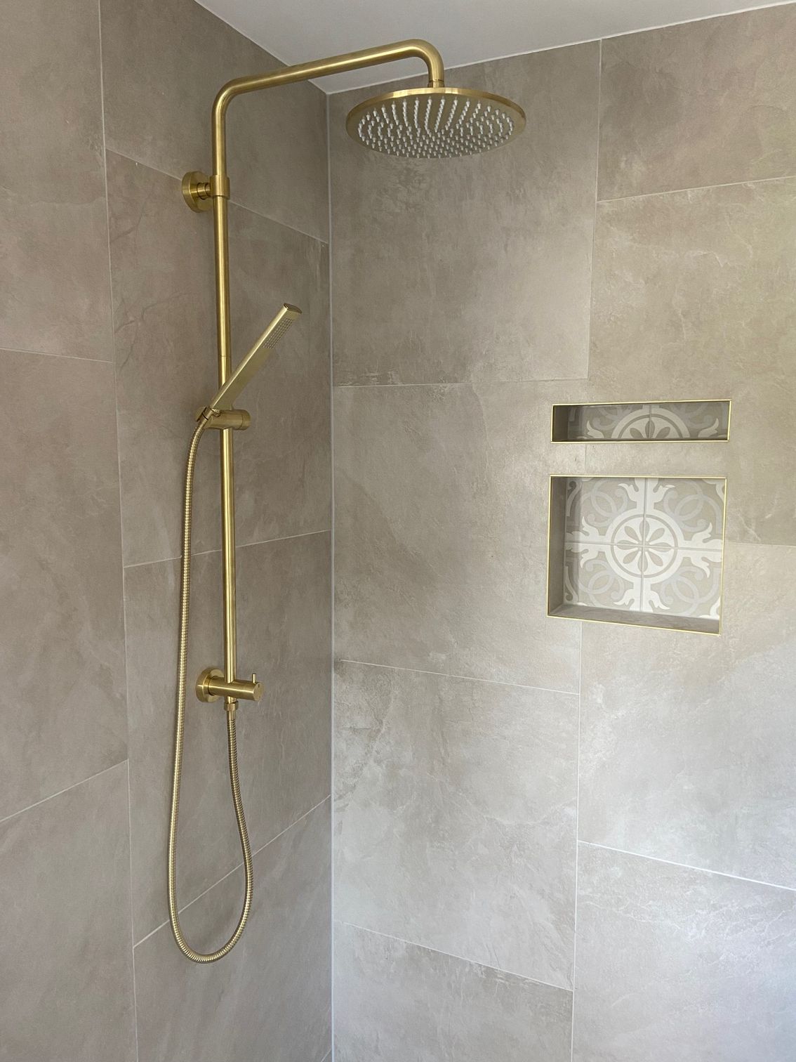 Gold showerhead and handheld shower fixtures against neutral tile, with built-in tiled shelf.