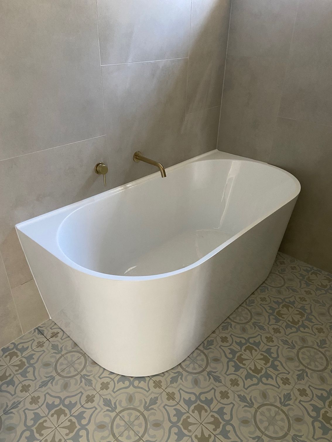 White freestanding bathtub in a modern bathroom with patterned tile floor and gold fixtures.