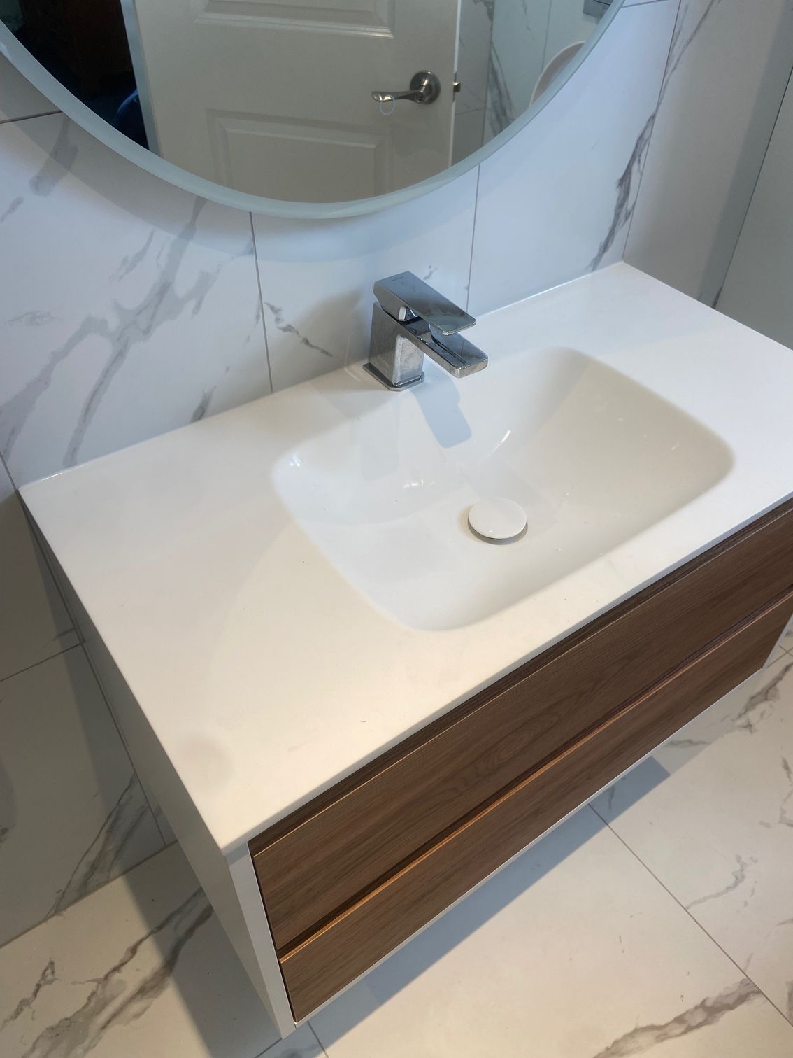 Bathroom vanity with white countertop, integrated sink, chrome faucet, and wood drawer against a marble-look wall.