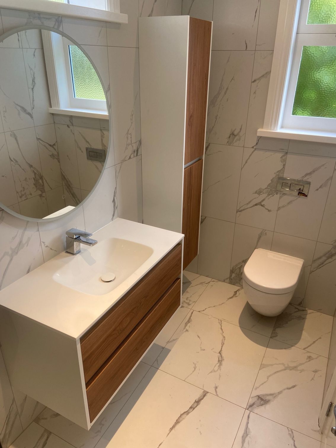 Modern bathroom with white marble tile, wood-toned vanity, and wall-mounted toilet.