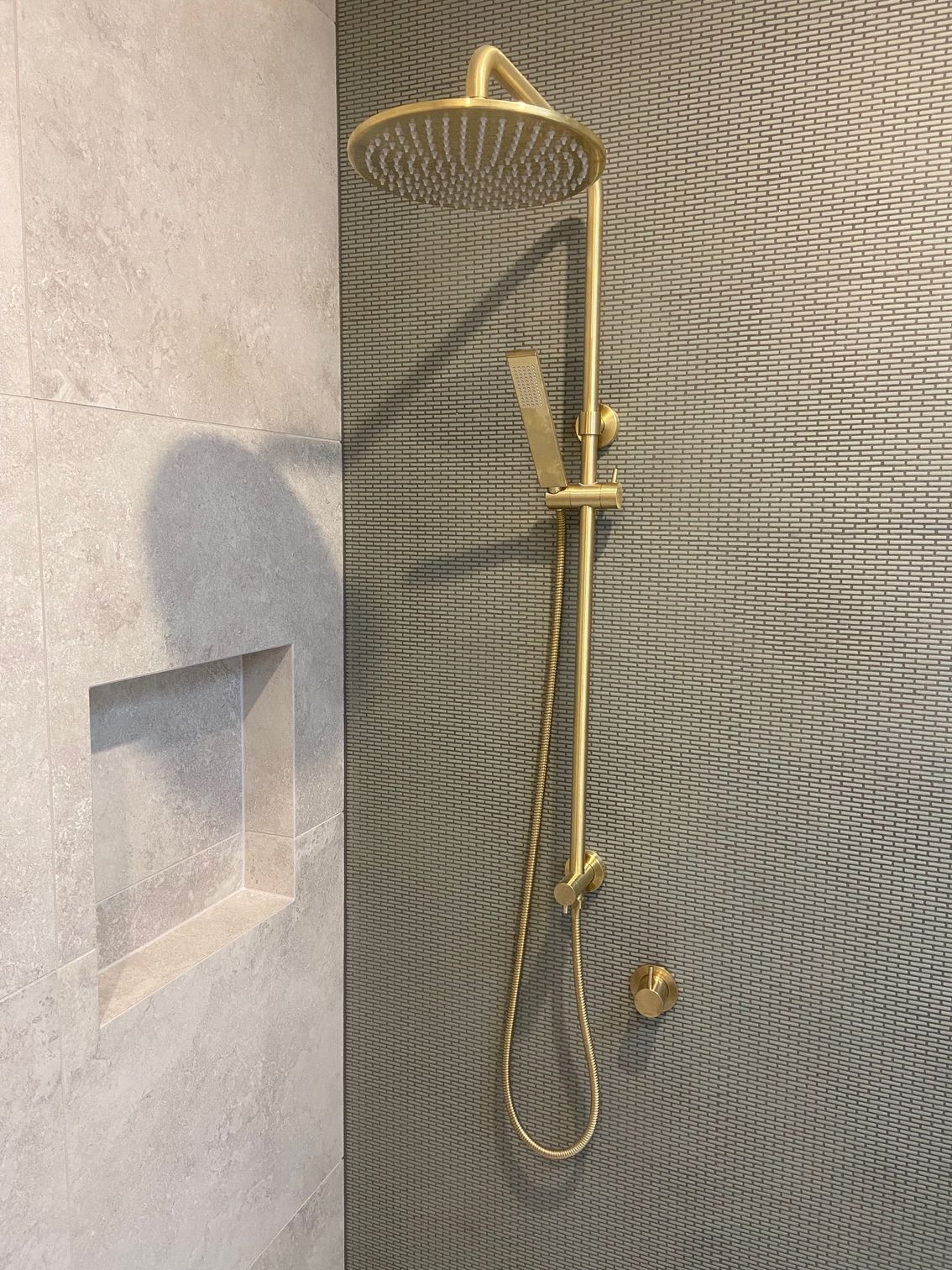 Gold shower head and handheld shower against a textured gray mosaic tile wall, next to a recessed alcove.
