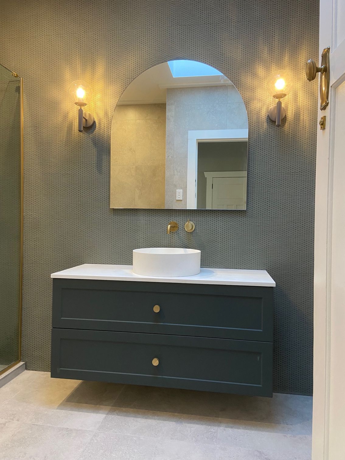 Bathroom with a dark teal floating vanity, round vessel sink, arched mirror, and mosaic tile wall.