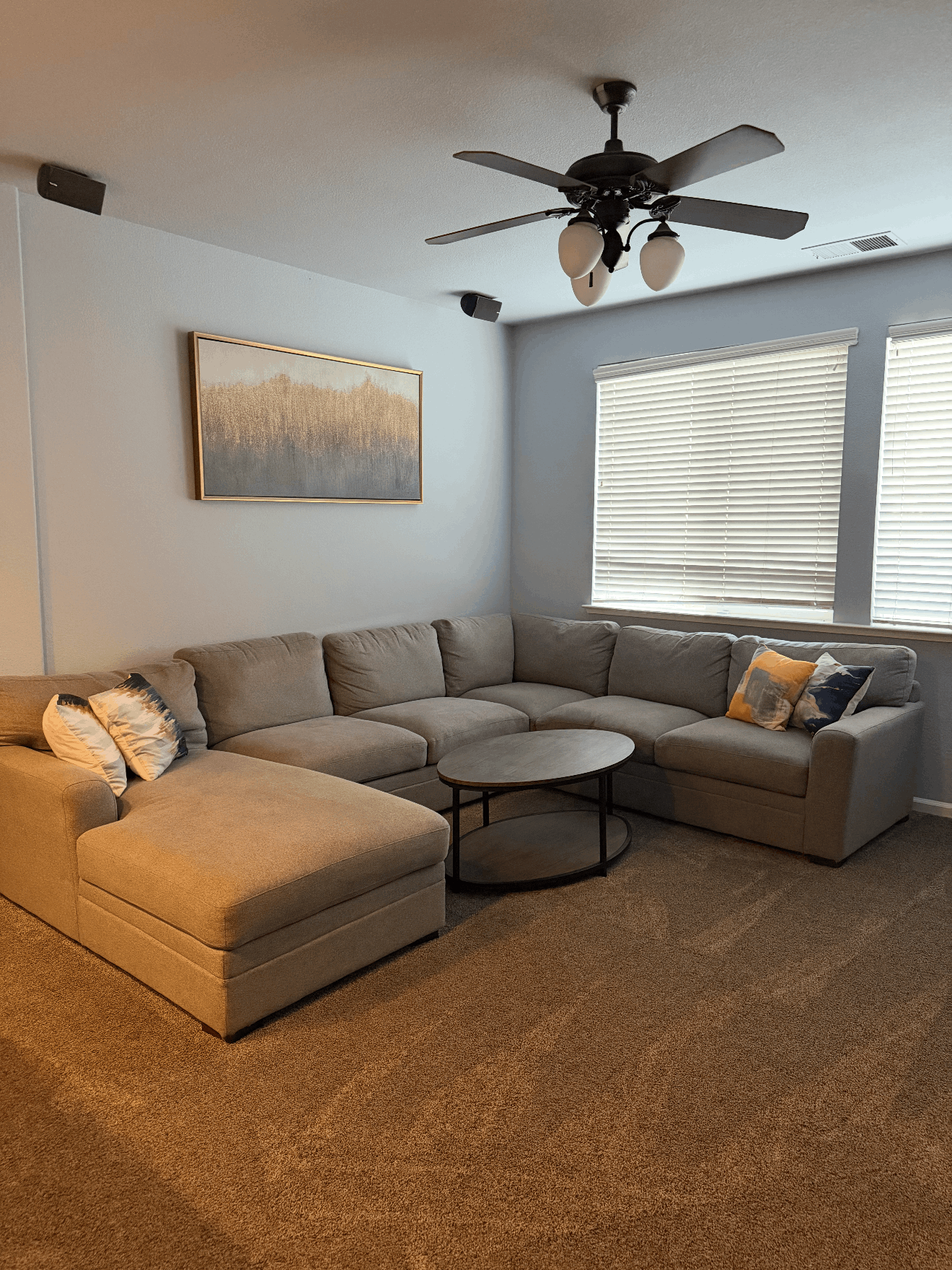Bright living room with gray sectional sofa, round coffee table, carpet, ceiling fan, and large window blinds.