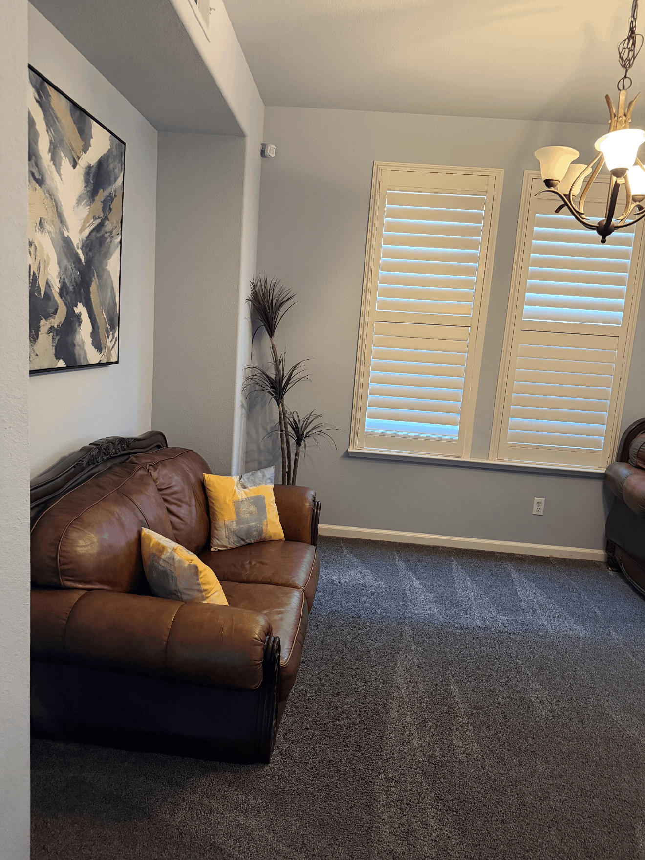 Living room with brown sofa, yellow pillows, gray carpet, window shutters, and chandelier.