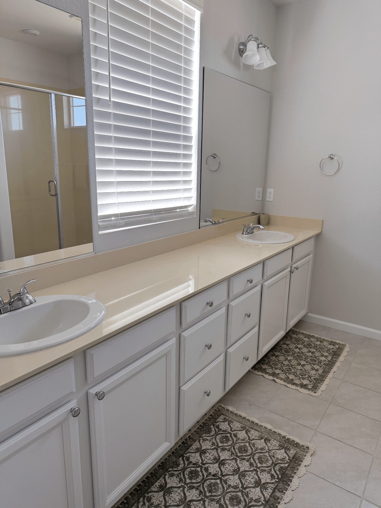 Bright white bathroom with double sinks, long countertop, cabinets, mirrors, and patterned rugs