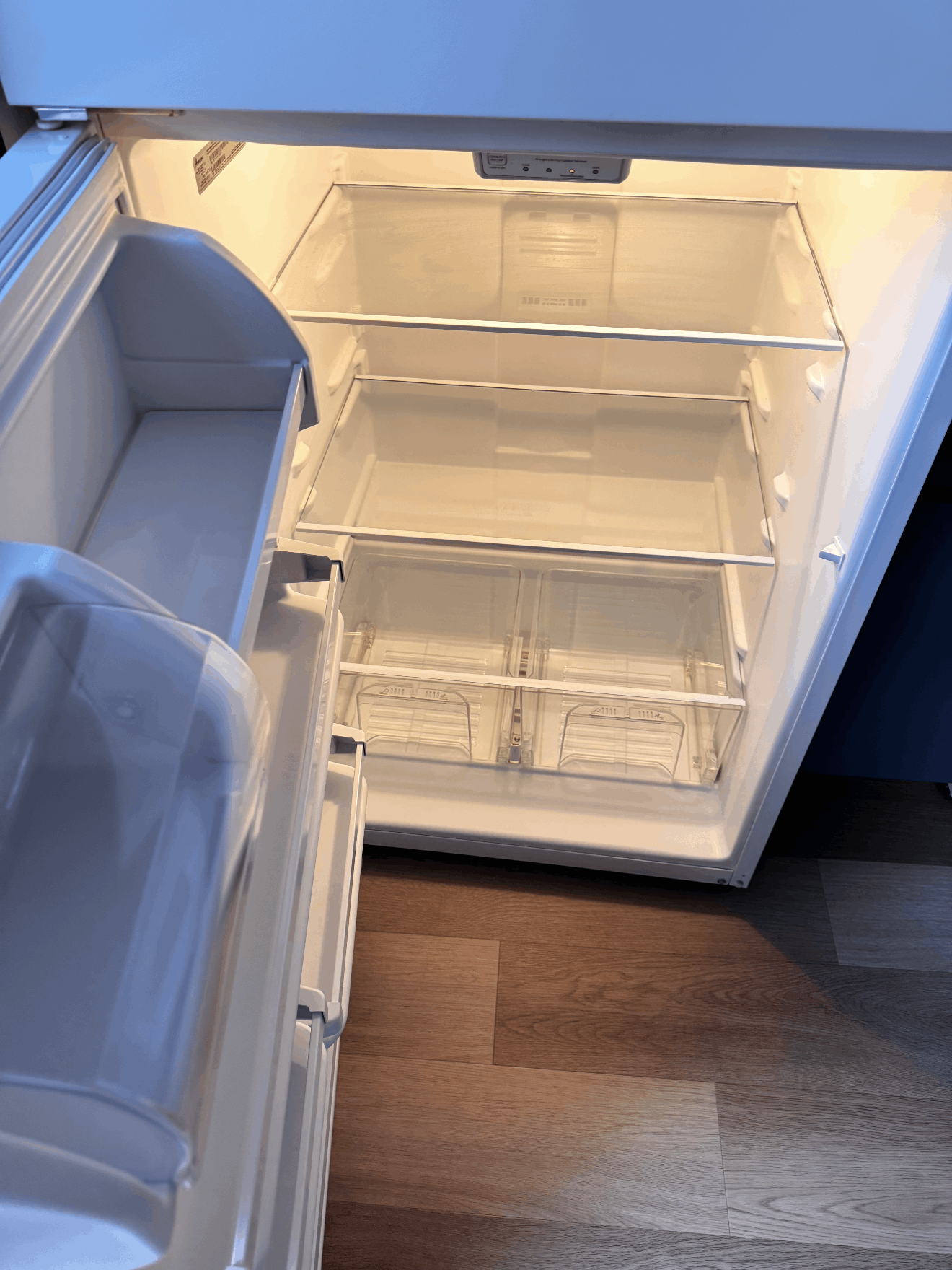 Open refrigerator with empty shelves and a door bin, lit from inside, beside a wooden floor.