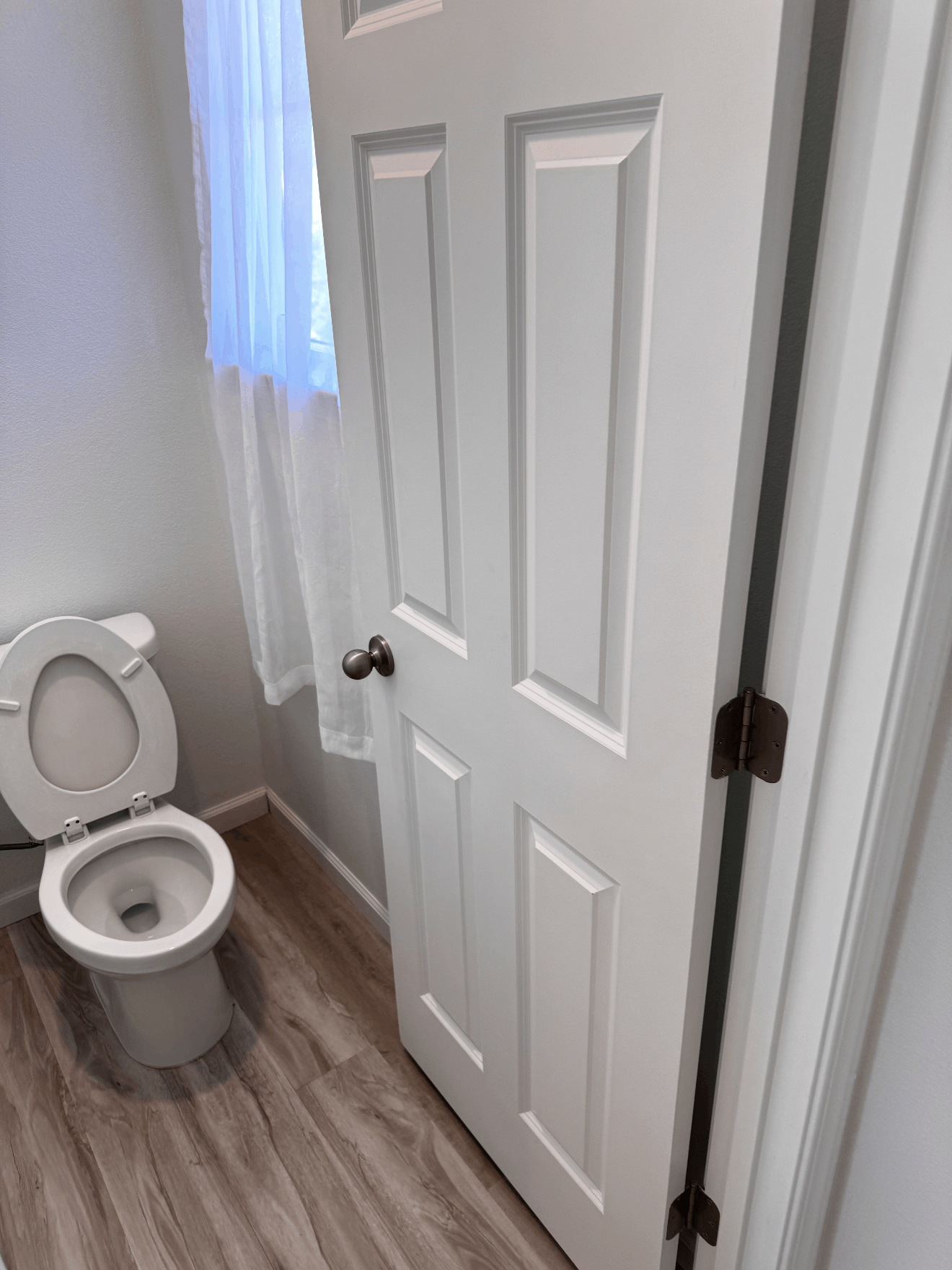 Bathroom with an open toilet, white door, and window with blue curtain.