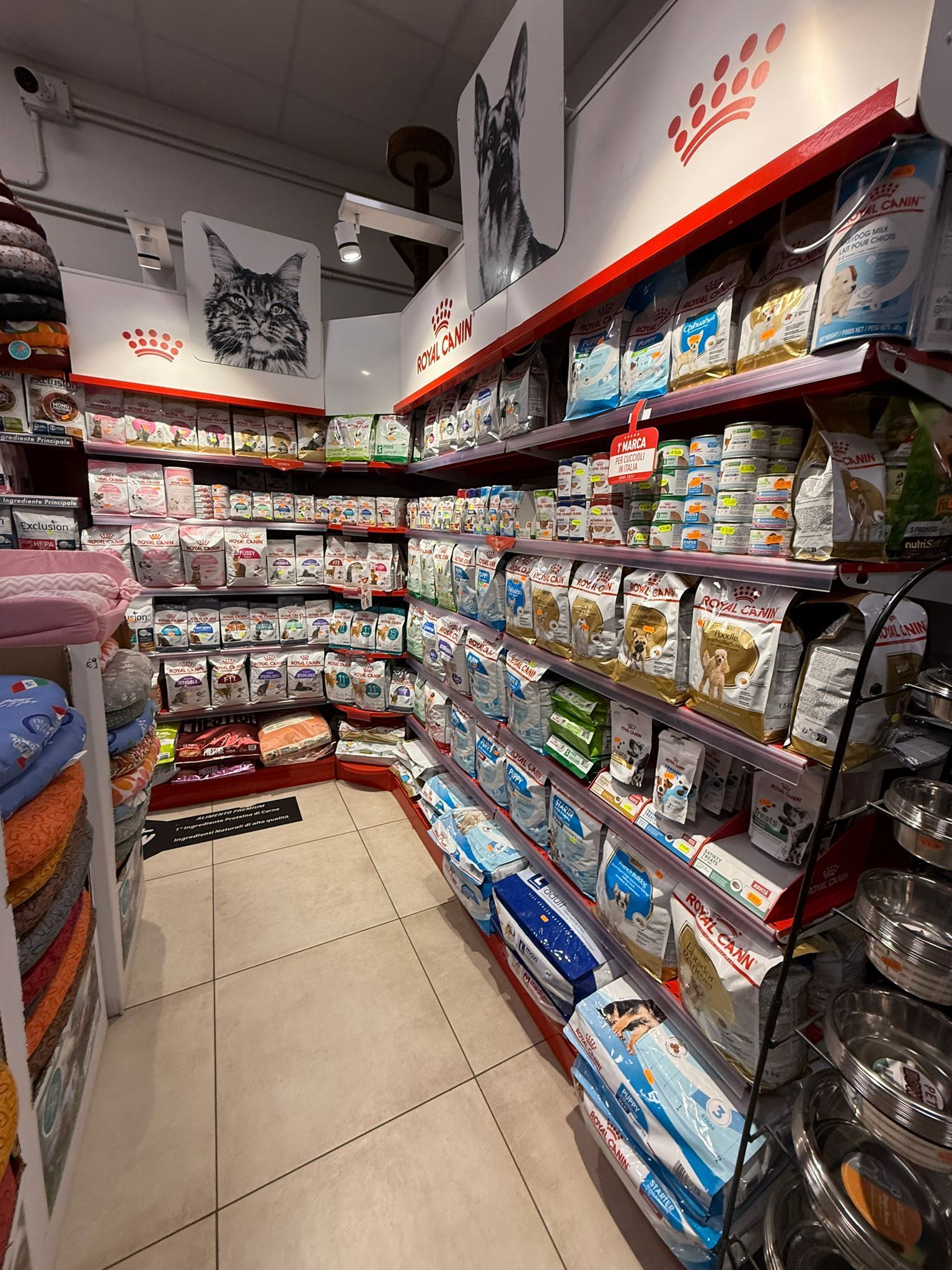 In un corridoio di un negozio di animali, scaffali metallici sono pieni di sacchetti e contenitori di cibo per animali, con cartelli che indicano cani e gatti.