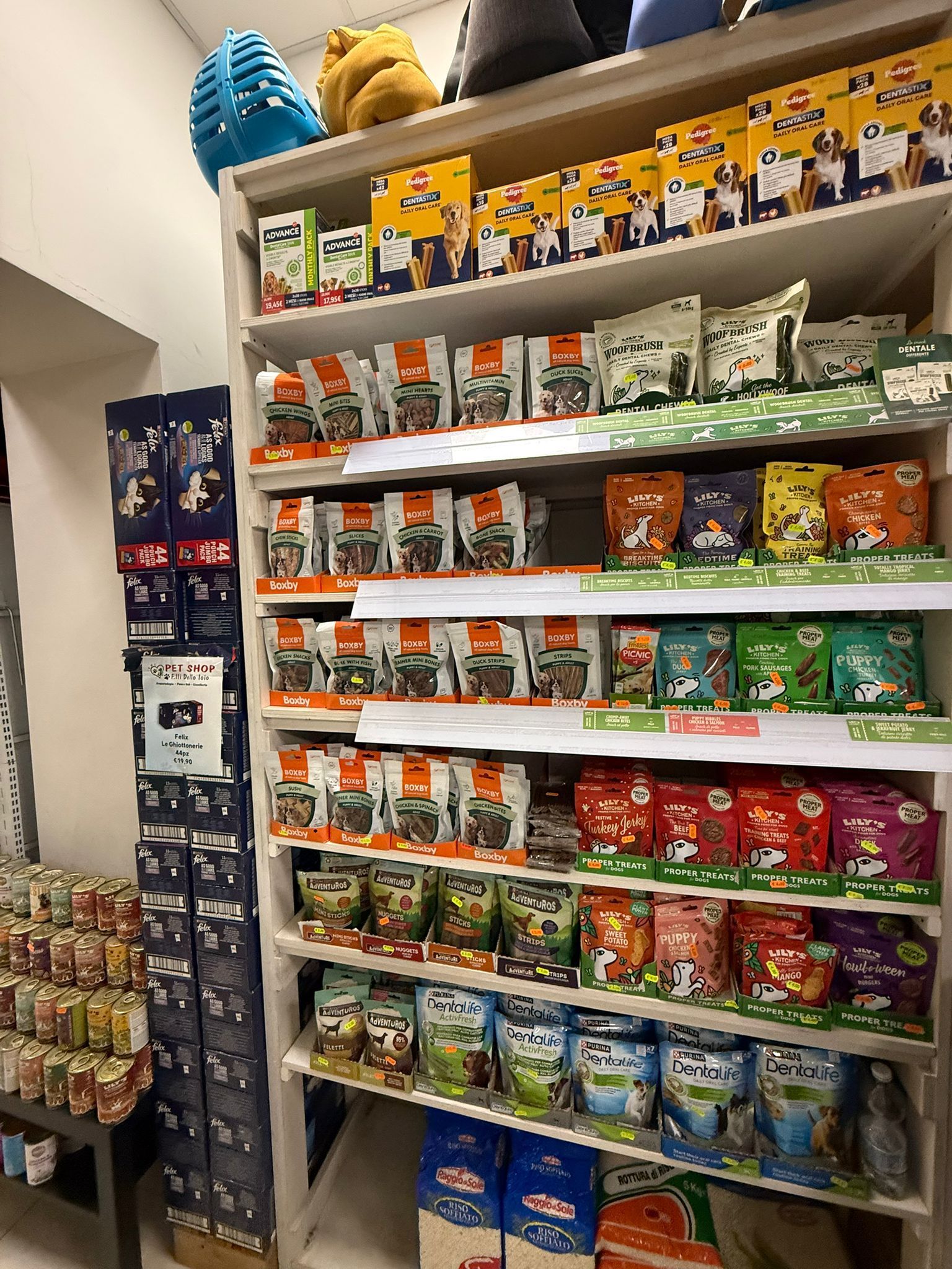 In un negozio, gli scaffali sono pieni di varie confezioni di cibo e snack per animali domestici, con un giocattolo blu sullo scaffale più alto.