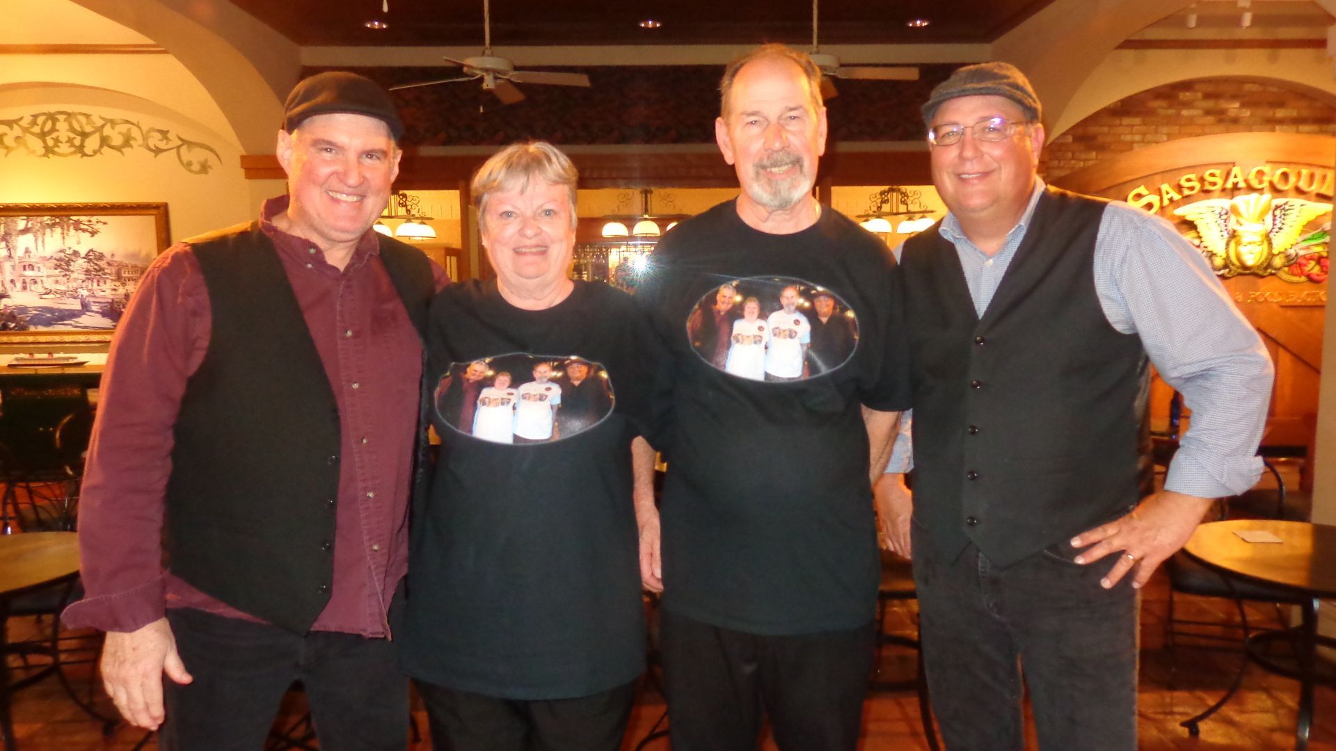Four people smiling, wearing matching black t-shirts and vests, posing in a restaurant with tables.