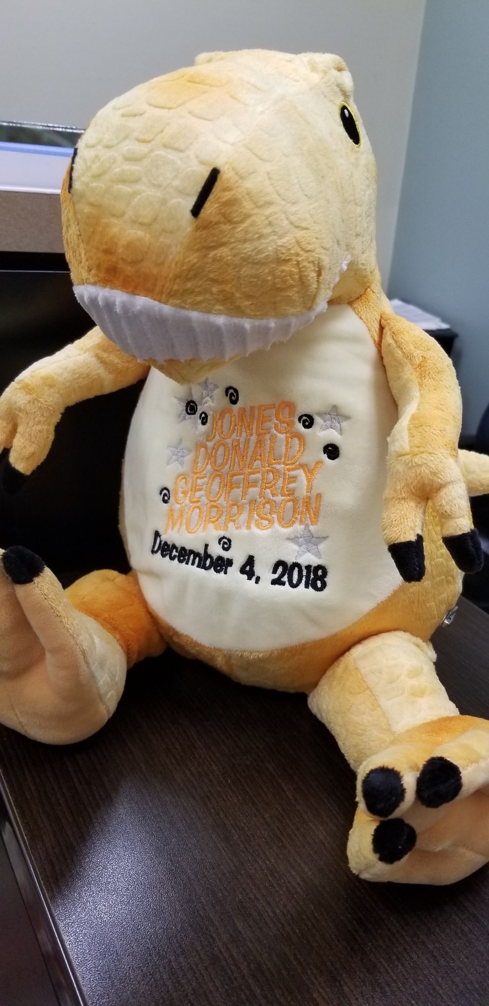 Yellow dinosaur stuffed animal with a personalized emblem on its belly, sitting on a dark surface.