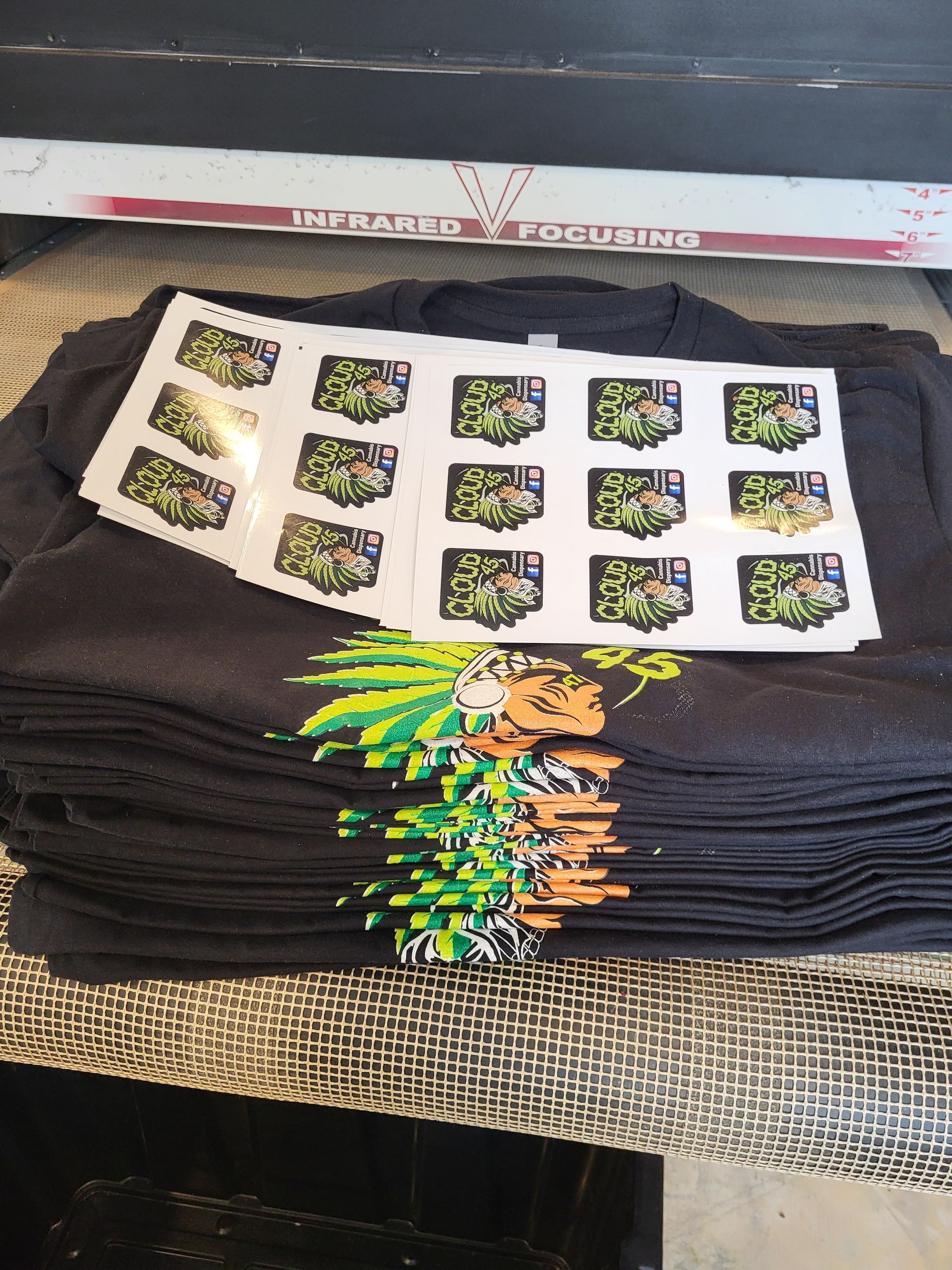 Pile of dark gray t-shirts with printed graphics, and sheets of stickers, on a textured surface.