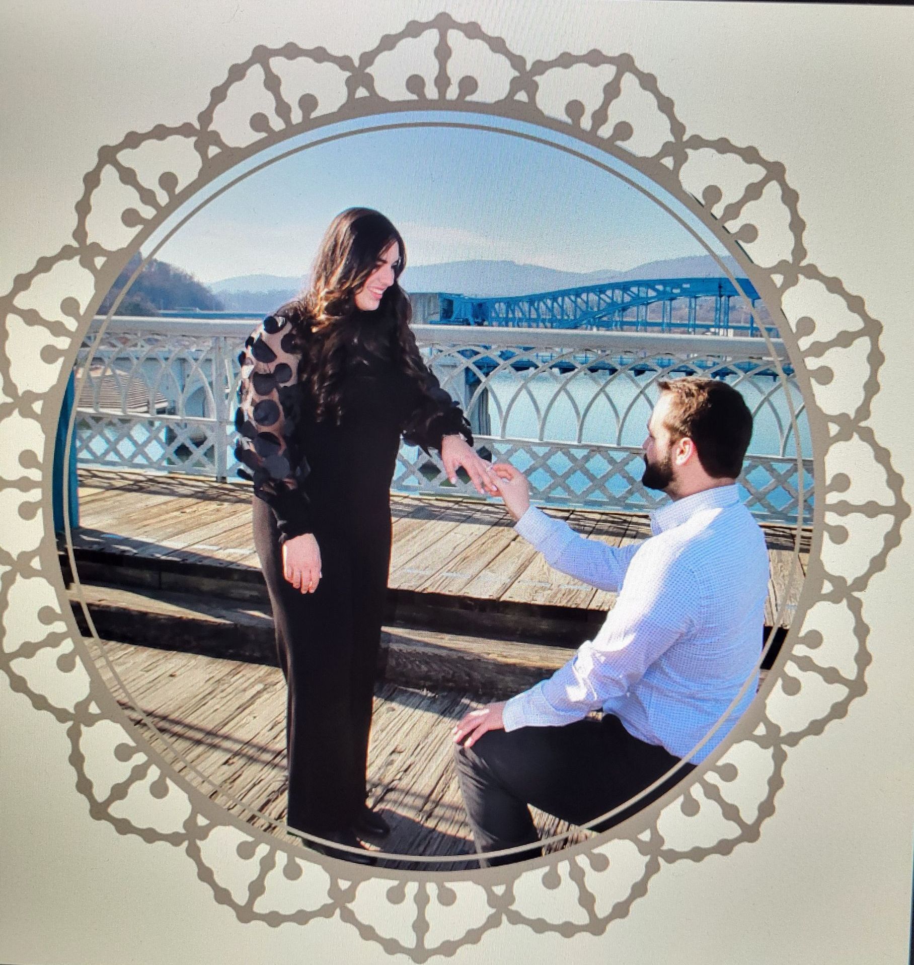 Man proposing to woman on a wooden pier with a bridge in the background. She is wearing a black dress.