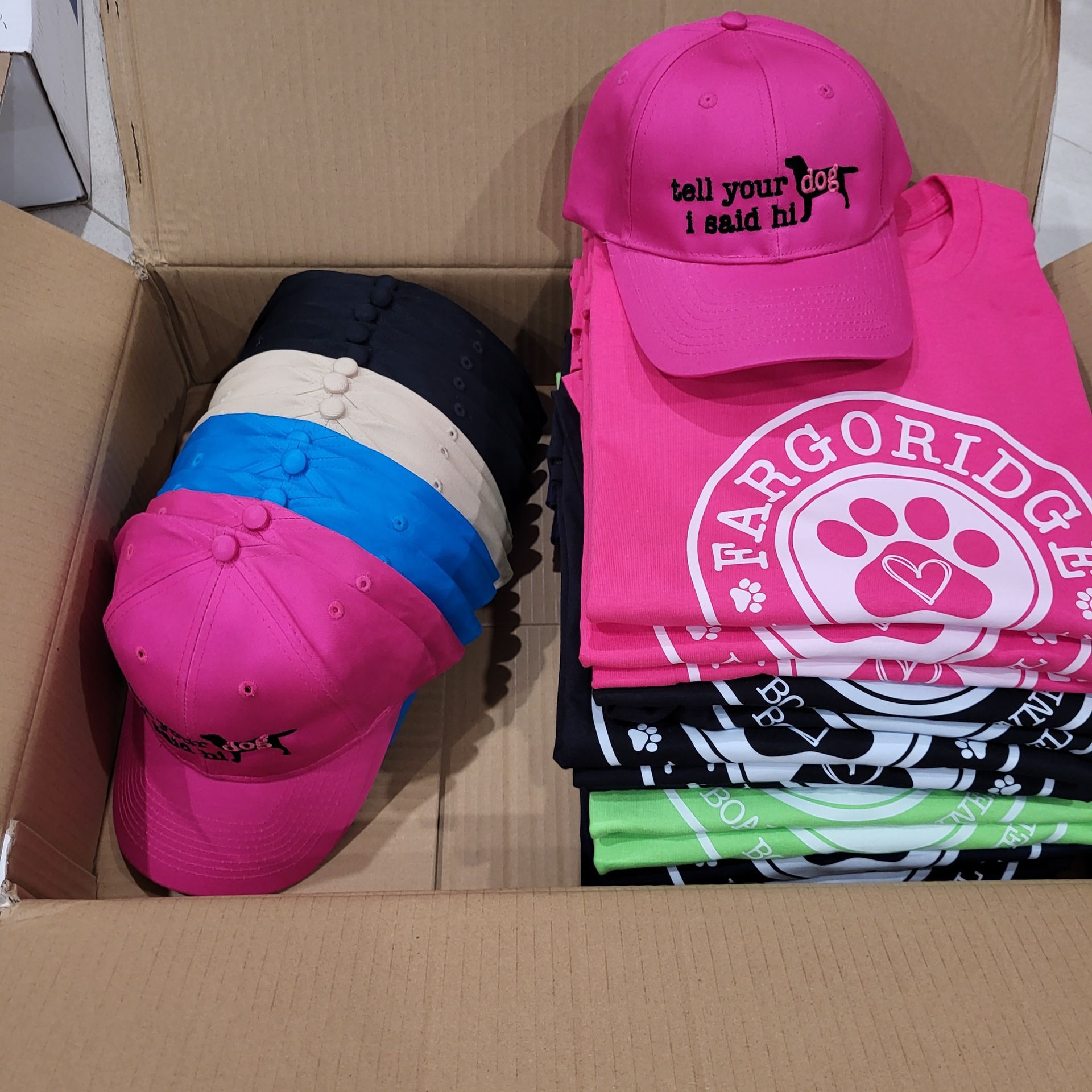 Cardboard box holding stacked t-shirts and caps; the shirts are in bright colors.
