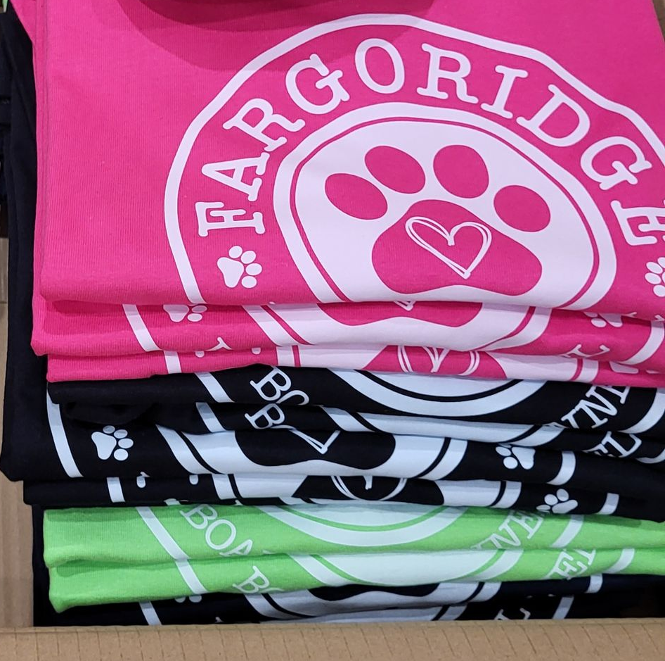 Stacked t-shirts in pink, white, black, and green, each with a Fargo Ridge pet paw logo.