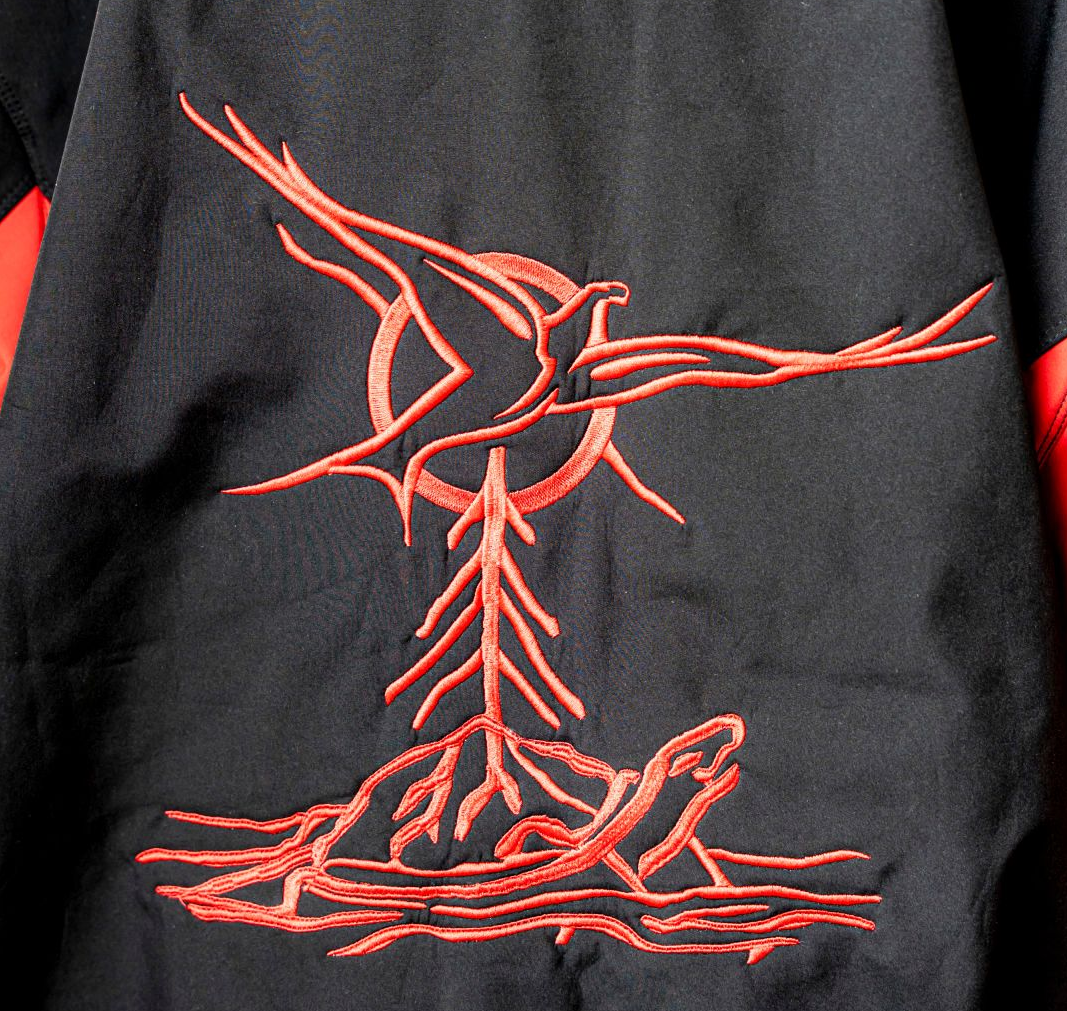 Embroidered red design on black jacket back: bird with outstretched wings above a tree-like form and a reclining turtle.