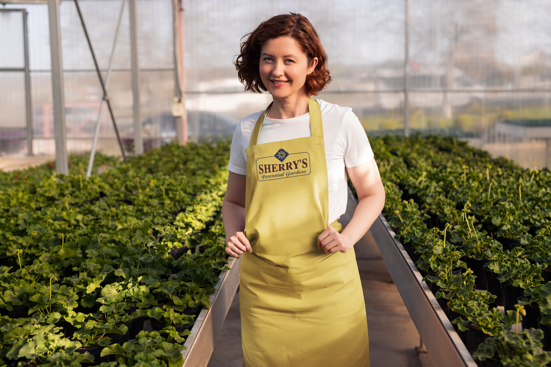 Woman in apron, smiling in a greenhouse, surrounded by plants.