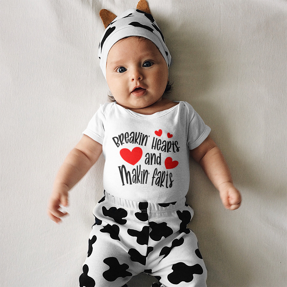 Baby in cow-themed outfit, wearing a cow hat, a onesie with hearts, and cow print pants, laying on a white surface.