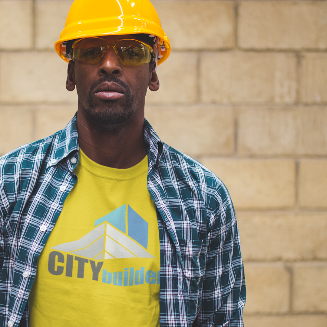 Construction worker wearing a yellow hard hat and safety glasses, standing in front of a brick wall.