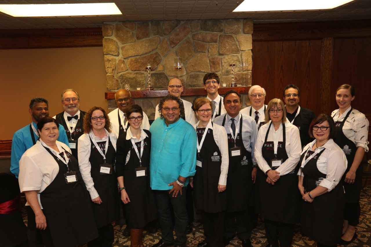 Group photo of people, some wearing aprons, gathered indoors by a stone fireplace.