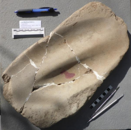 Broken beige stone artifact with white cracks, photographed next to a pen and scale ruler.