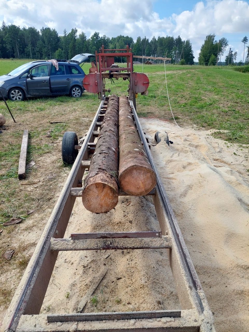 Logs on a sawmill track outdoors, with a blue car and red machinery in the background.