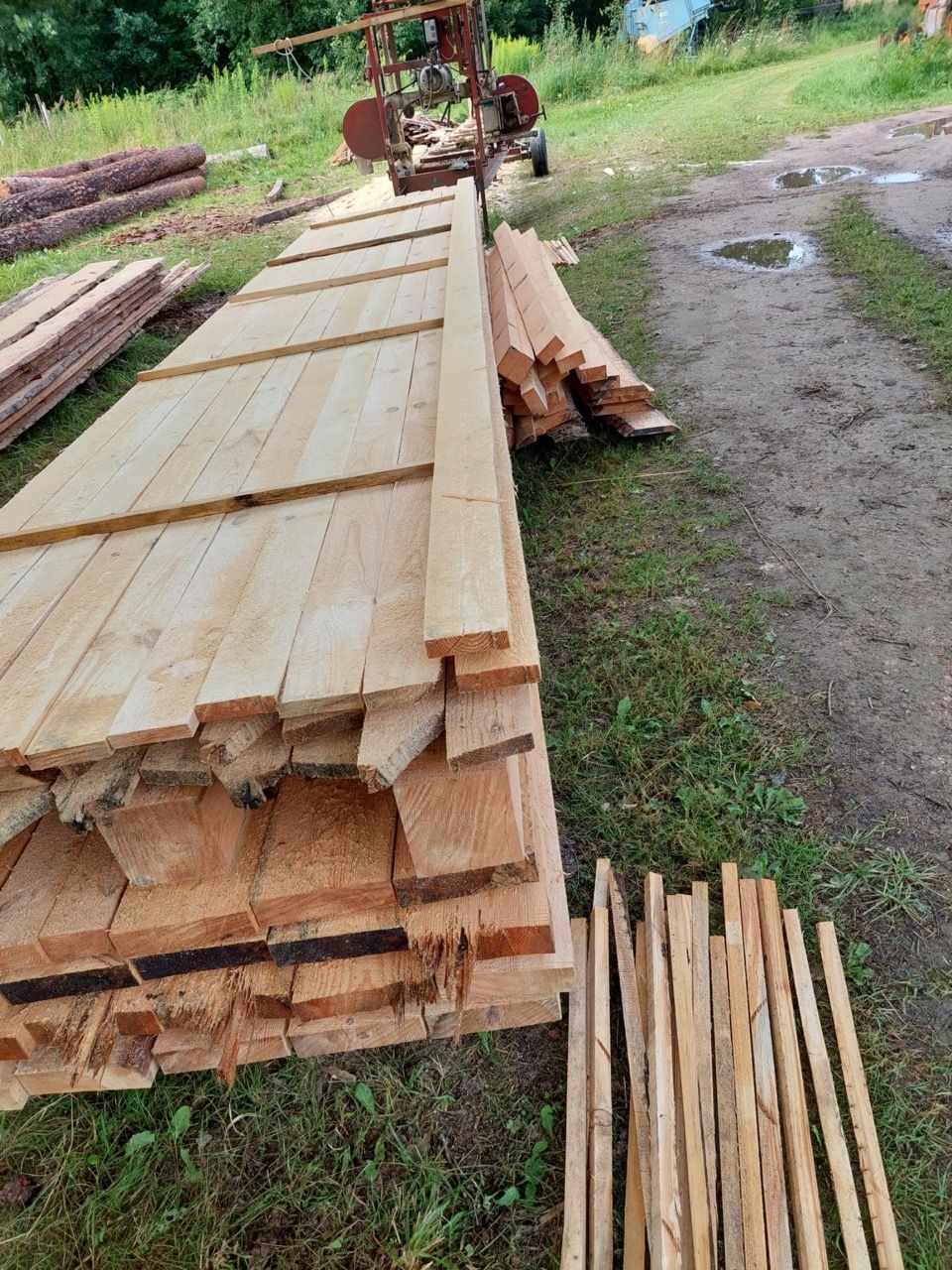 Stacked wooden planks drying outdoors beside a dirt path, with a small pile of sticks in front.