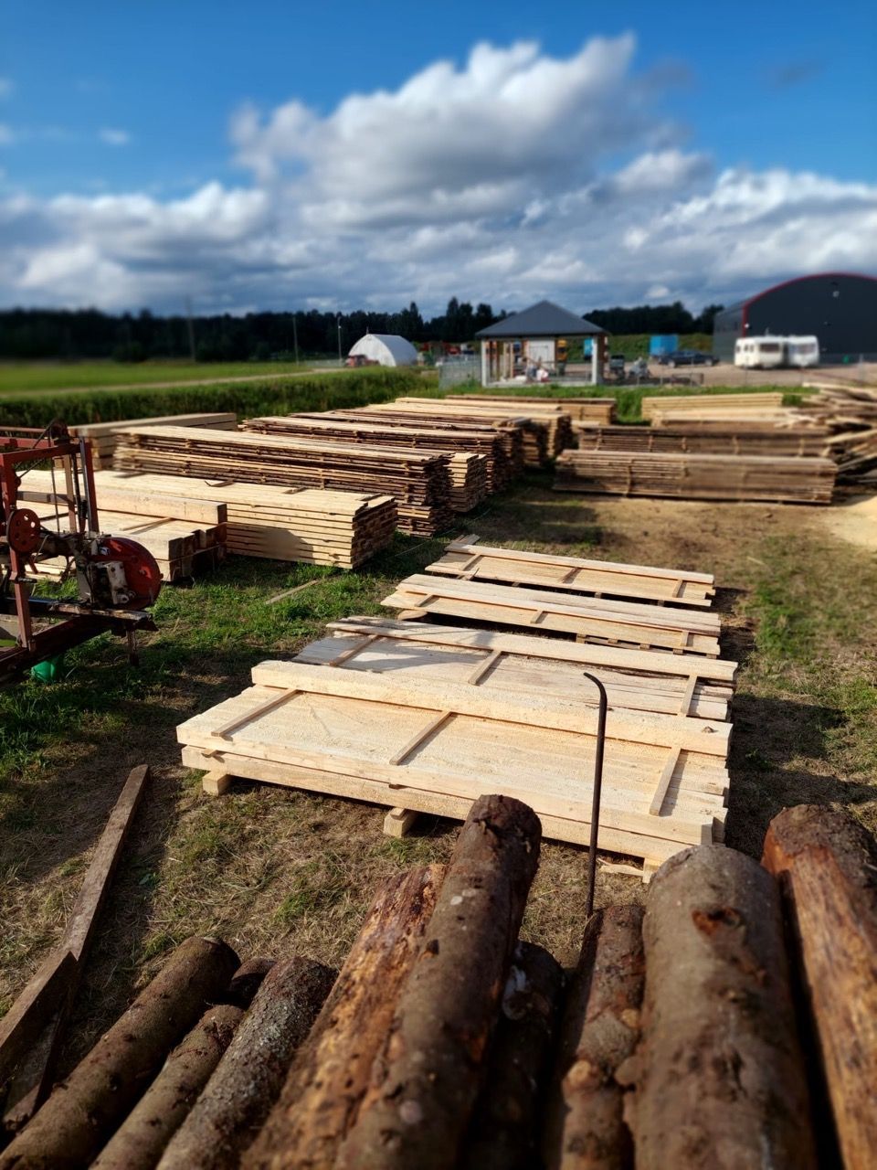 Stacked lumber and logs in a rural field under a cloudy sky, with a house in the background