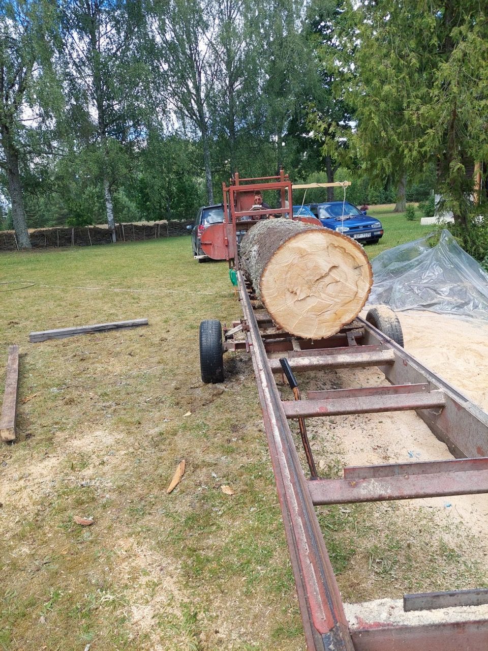 Log splitter trailer with a large log, parked on grass near trees and wood stacks