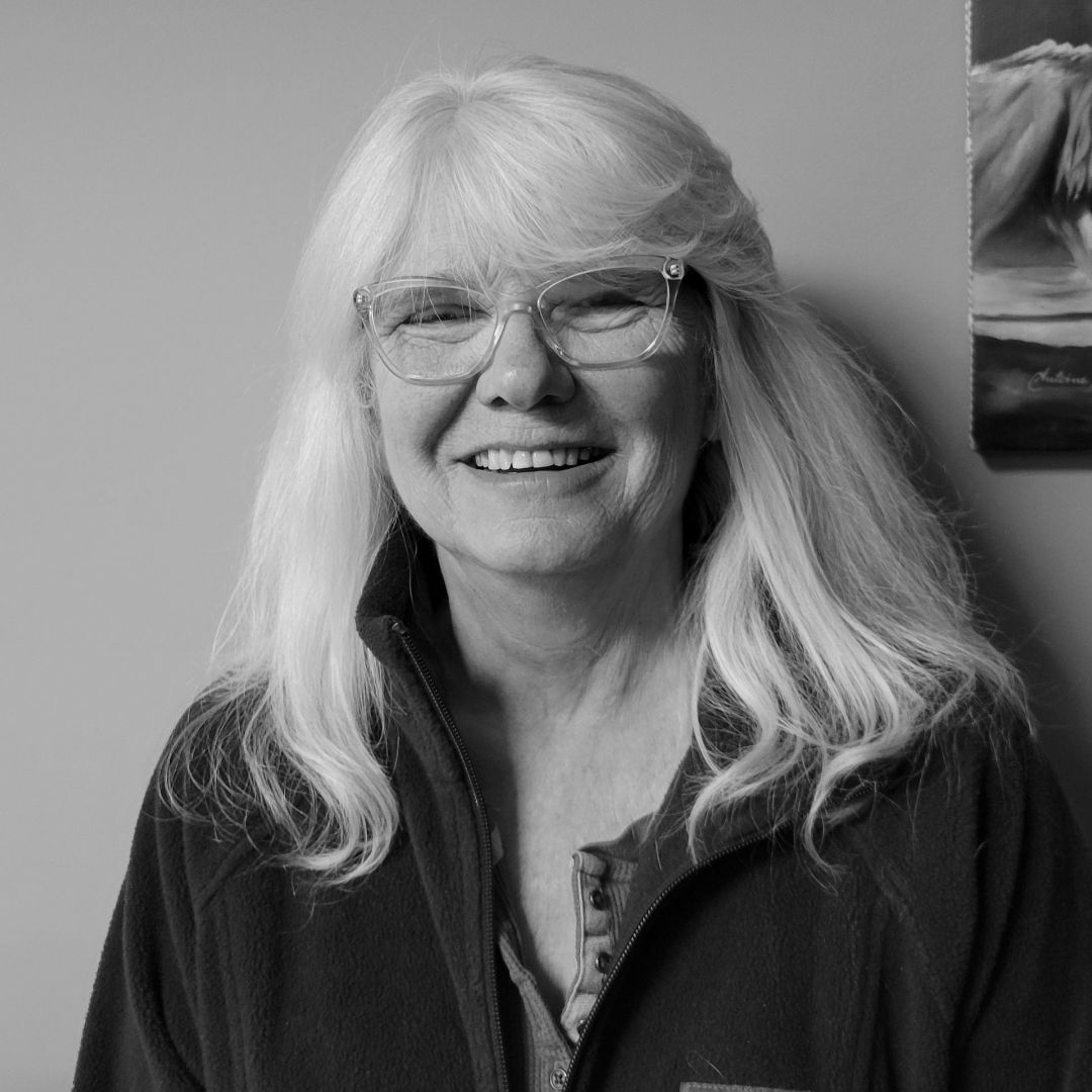 A woman wearing glasses and a black jacket is smiling in a black and white photo.