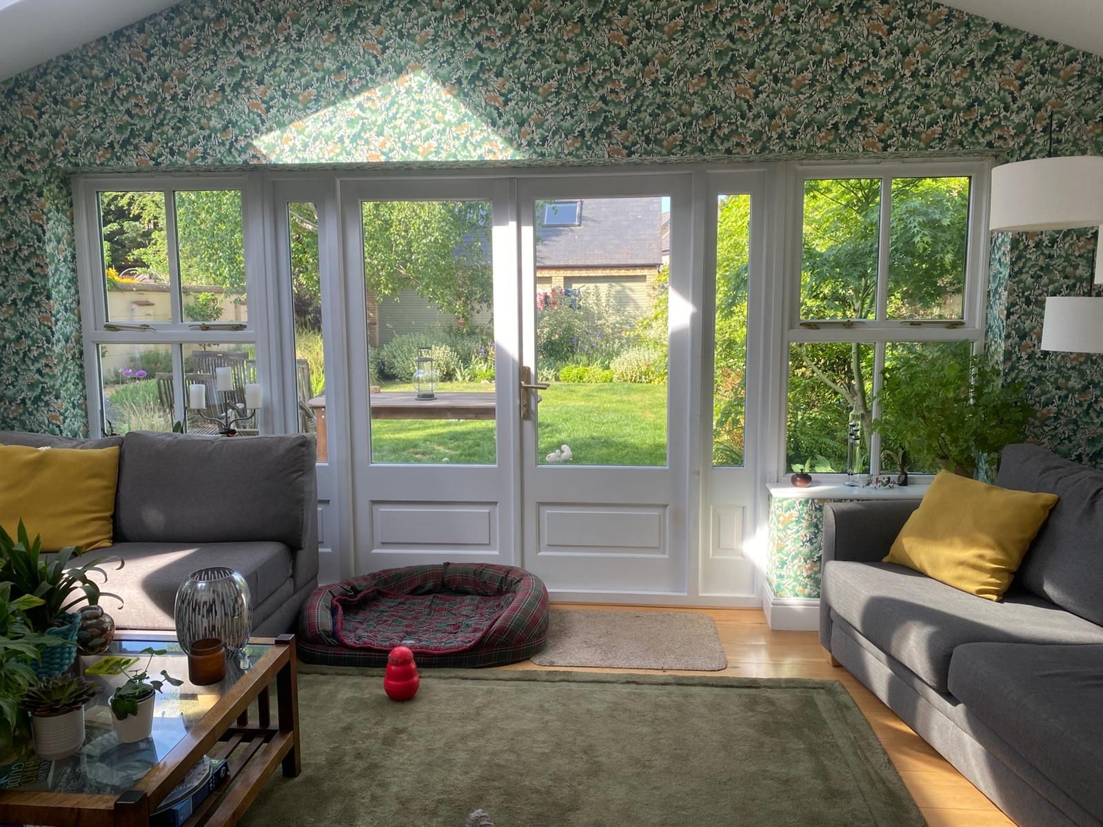 Bright living room with sofa, coffee table, green rug, and timber framed French doors opening to a sunny garden.