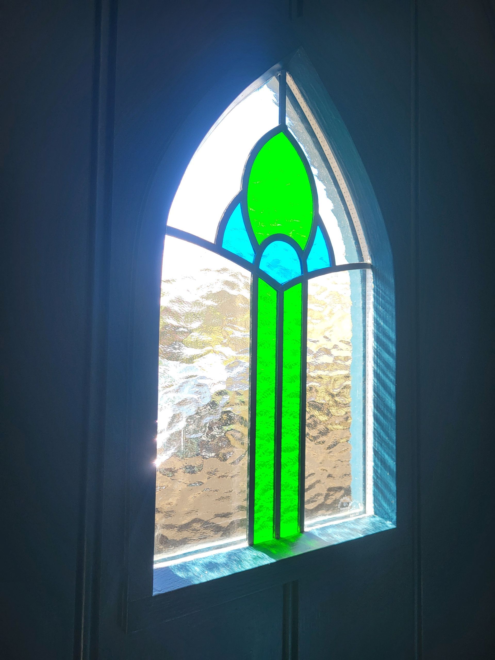 Green and blue stained-glass window in an arched opening, glowing with daylight.