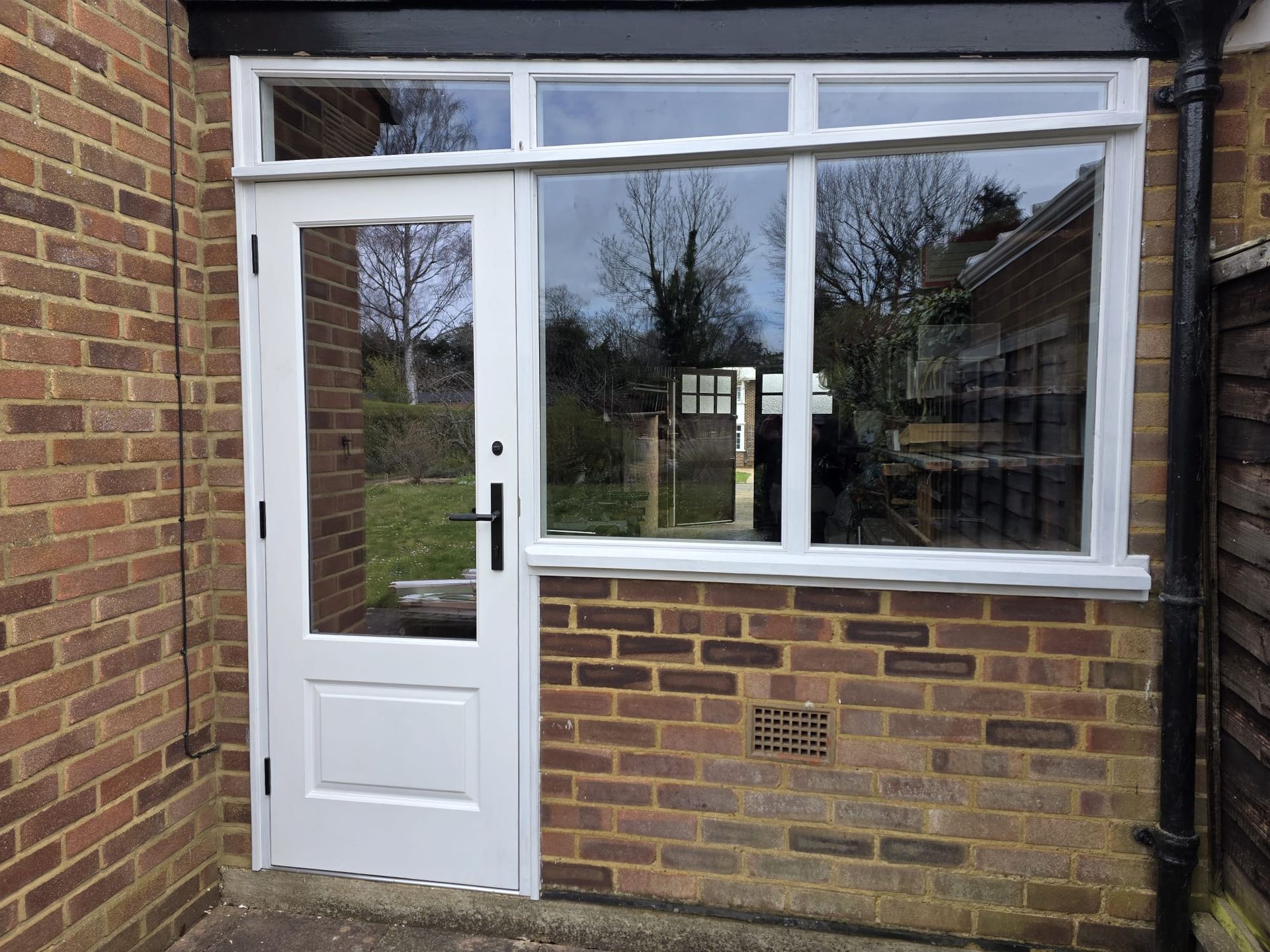 Brick house entrance with wooden framed door, large window, and black downspout.