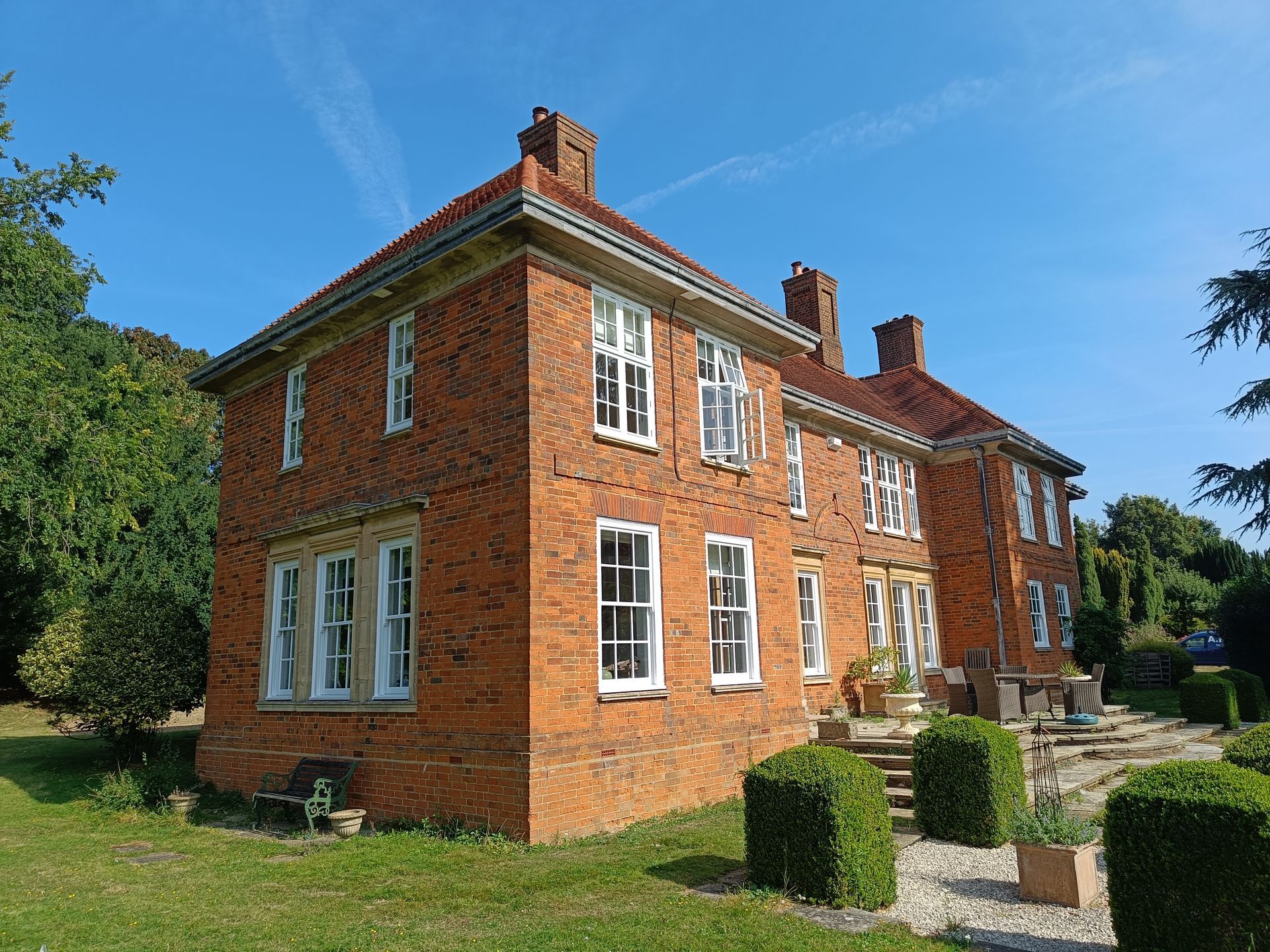 Red brick listed country house with white-timber replacement sliding sash windows and chimneys, surrounded by a garden on a sunny day