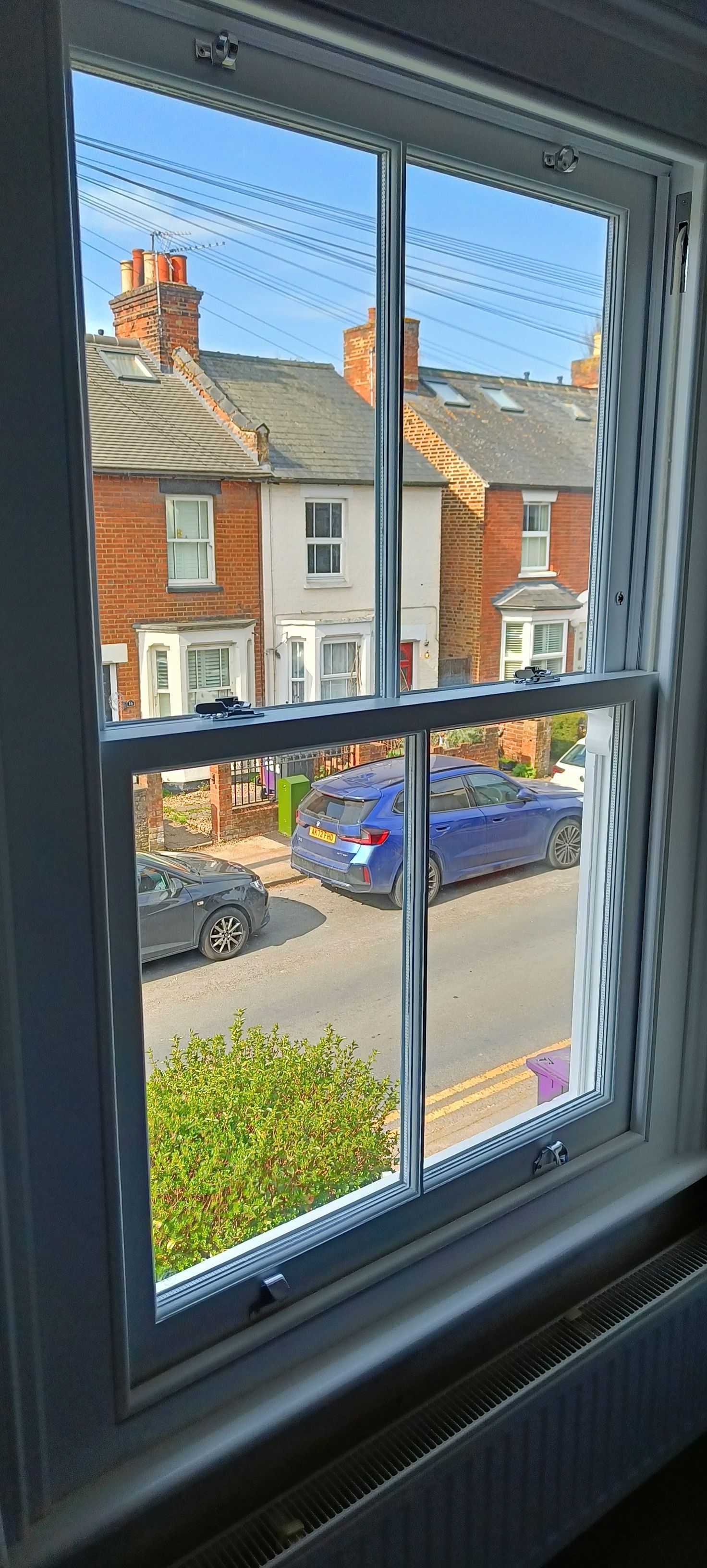 View through wooden manufactured sliding sash window of a residential street with brick houses, a parked blue car, and a tree.