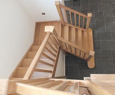 Wooden bespoke manufactured staircase viewed from above, with a central landing and dark tiled floor below.