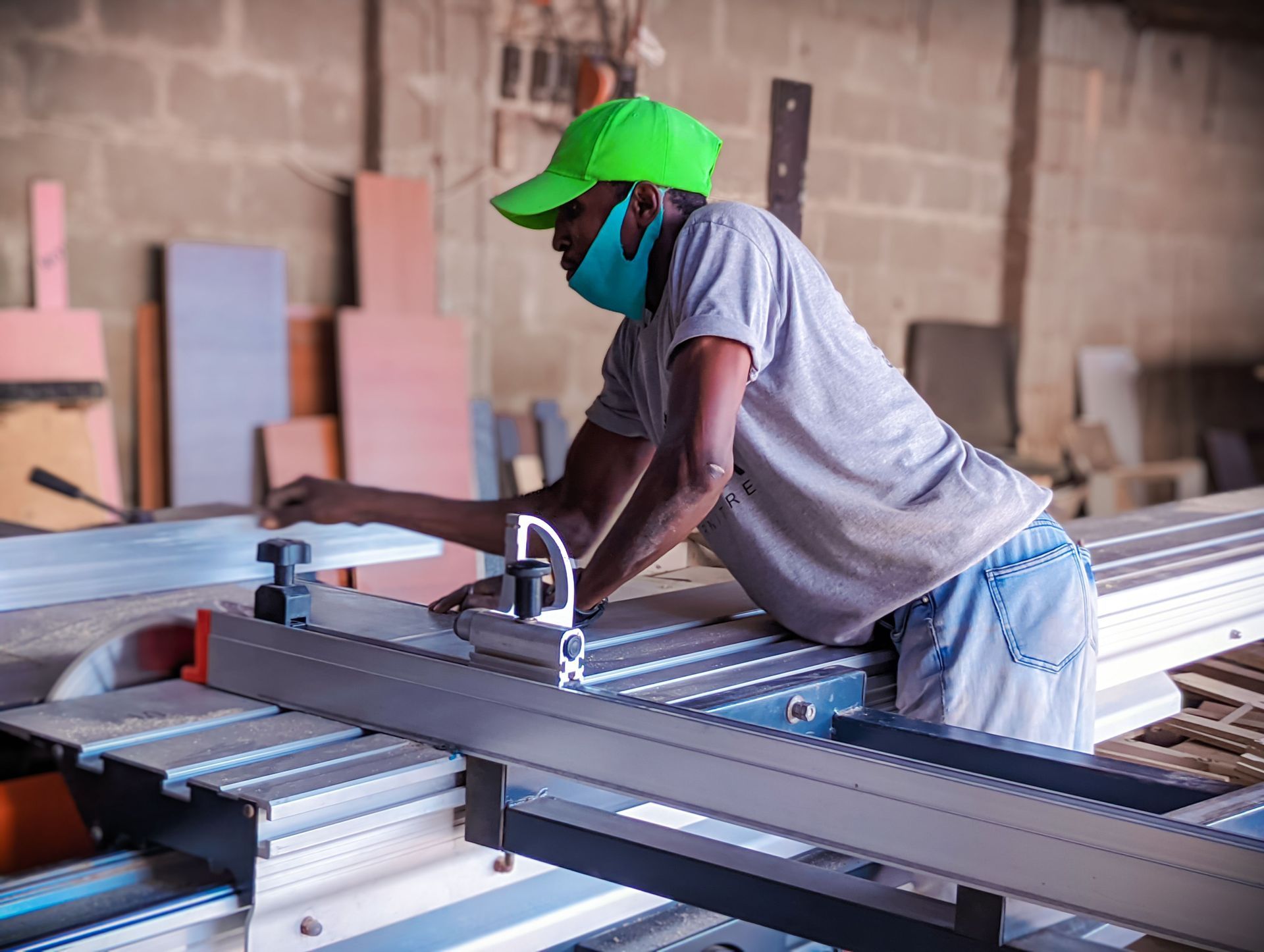 Worker in a green cap operating a table saw in a workshop with lumber stacks.