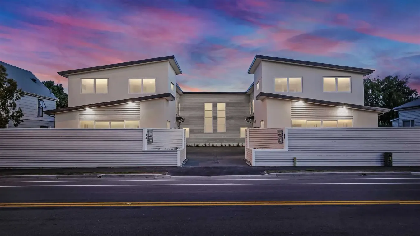 Modern two-story house at dusk with lit windows, a white fence, and a paved street in front