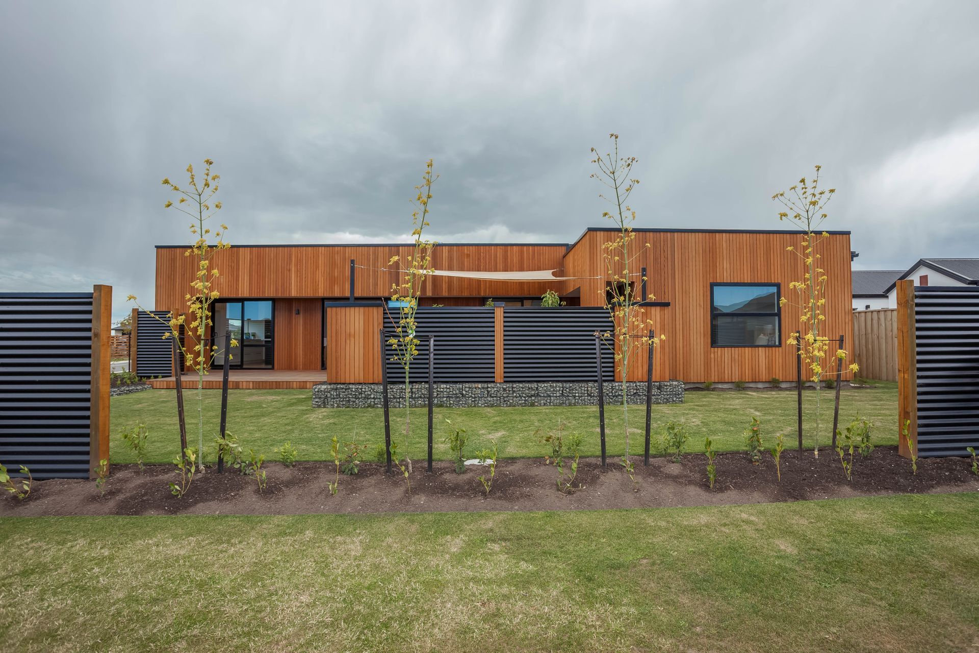 Modern wooden house with black accents behind a fenced front garden under a cloudy sky
