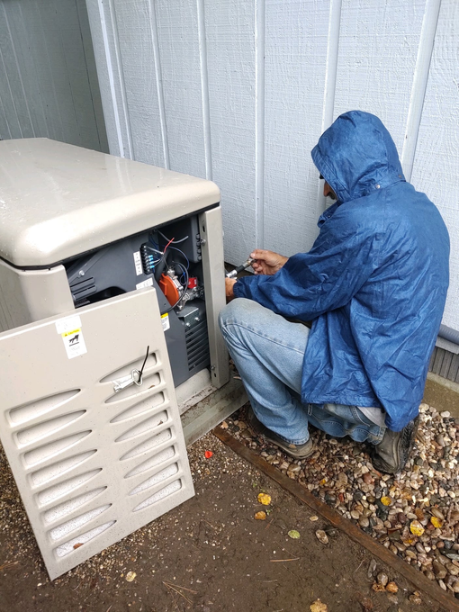 Person in a blue rain jacket kneels by outdoor electrical equipment beside a building.