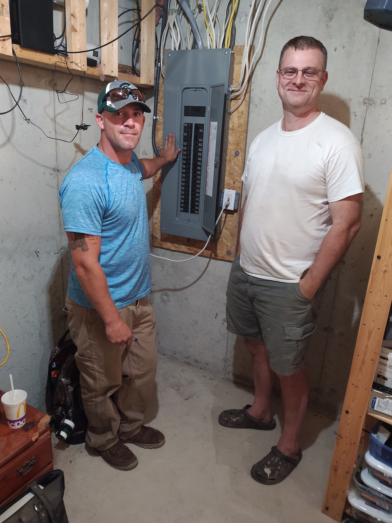 Two men standing beside an open electrical panel in a basement with exposed wiring