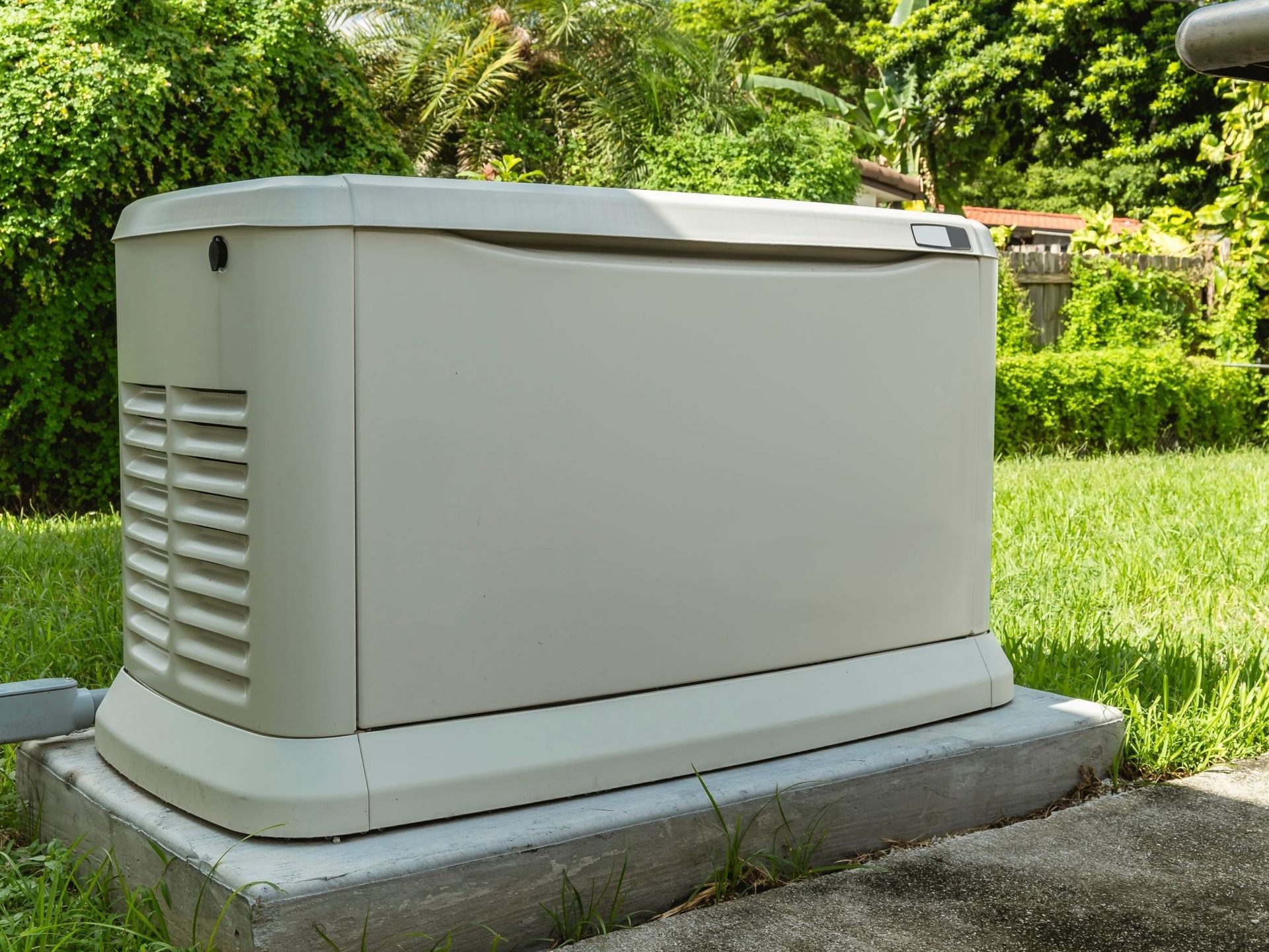 Outdoor beige backup generator on a concrete pad beside a grassy yard and trees