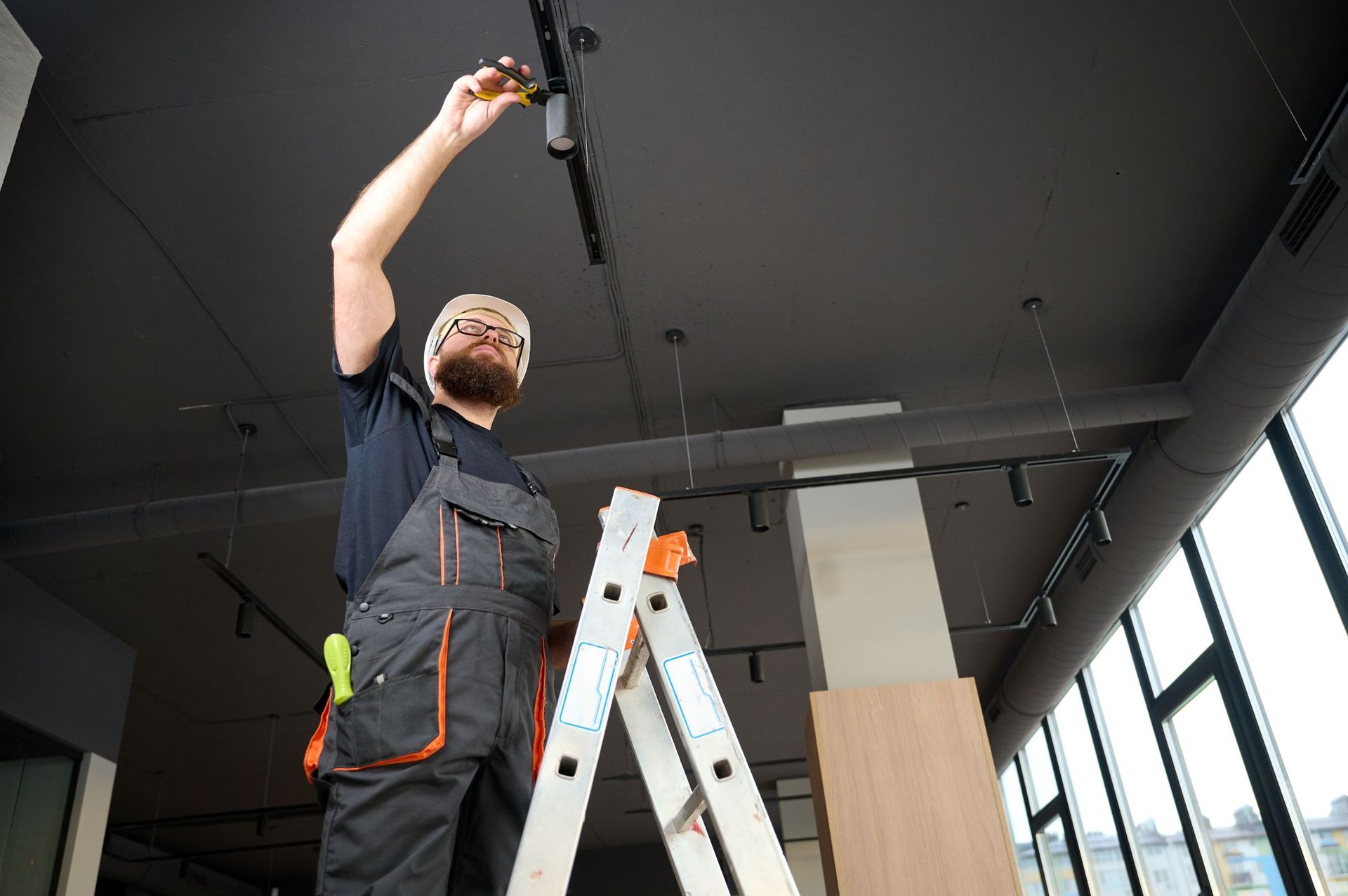 Worker on a ladder installing a black ceiling fixture in a modern room.