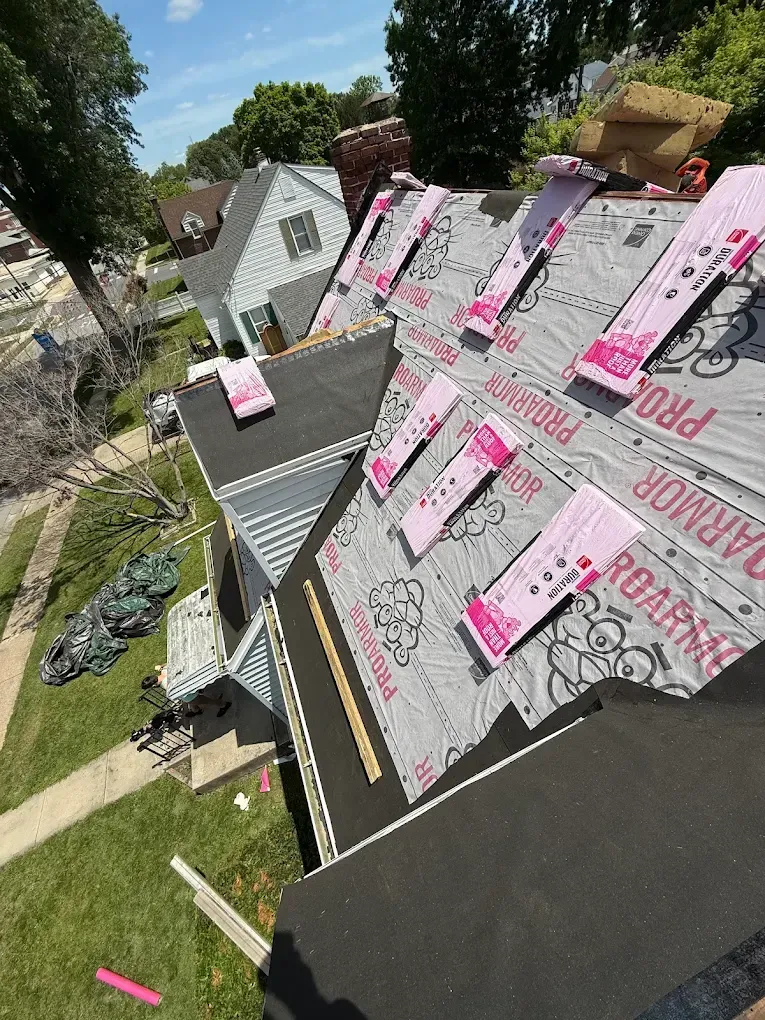 Roof under construction with pink roofing underlayment and shingles stacked on a sloped house roof.