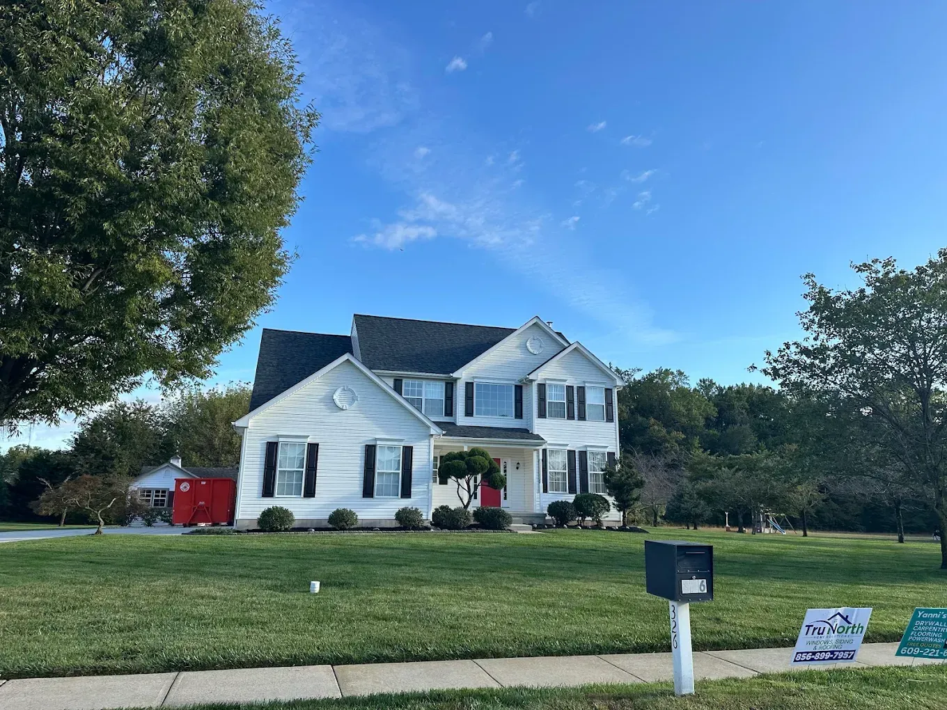 Two-story white house with blue roof and red flower pots on a sunny suburban lawn