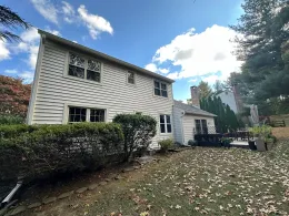 Two-story white house with black shutters, front yard covered in fallen leaves under a cloudy blue sky